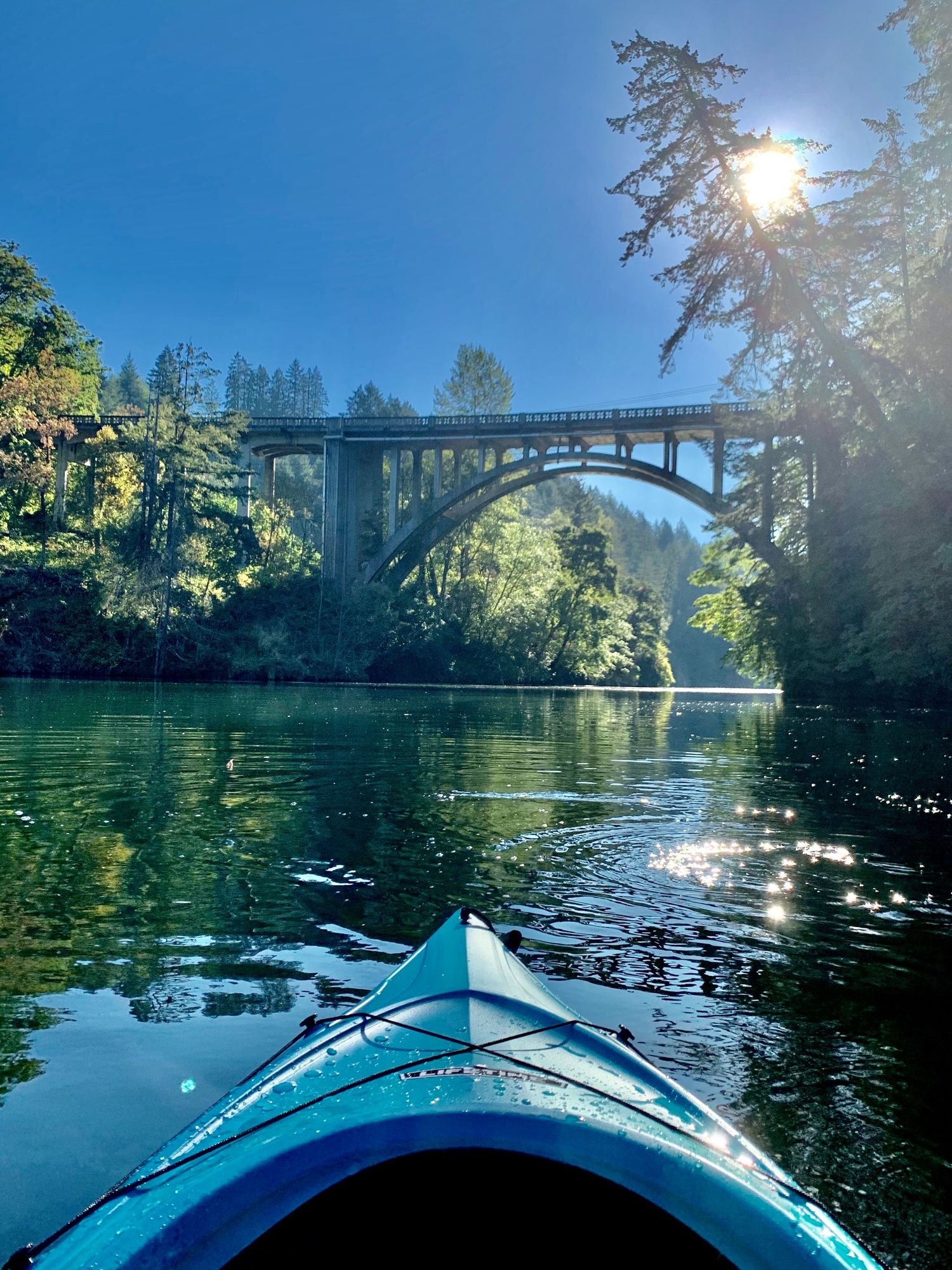 Blue kayak bow glides toward scenic stone arch bridge surrounded by tall evergreen trees on calm river.
