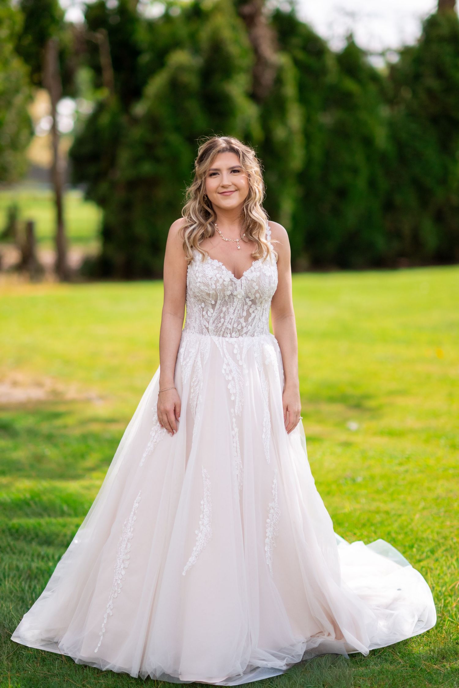 Beautiful bride in floral lace A-line wedding gown stands smiling on green lawn with evergreen trees behind.
