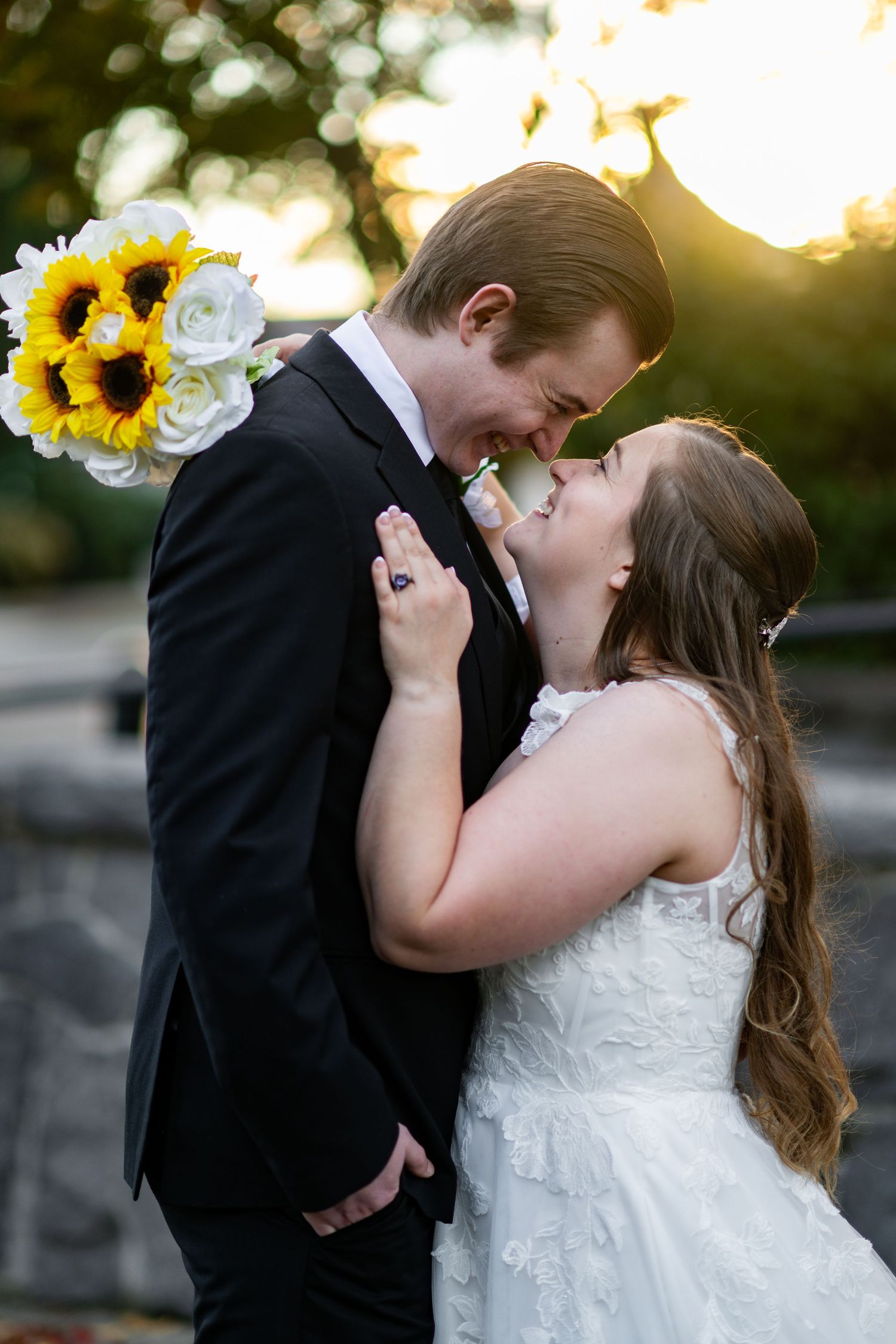 Bride and groom share romantic sunset moment with sunflower and white rose bouquet at outdoor wedding venue.