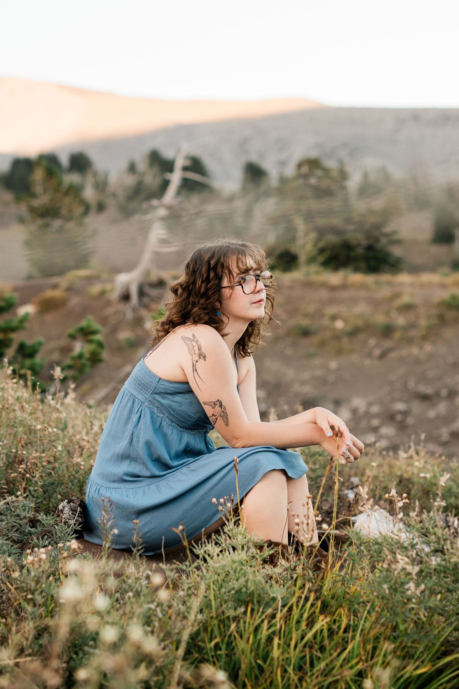 Person in blue dress sits thoughtfully in a field of wildflowers during golden hour.
