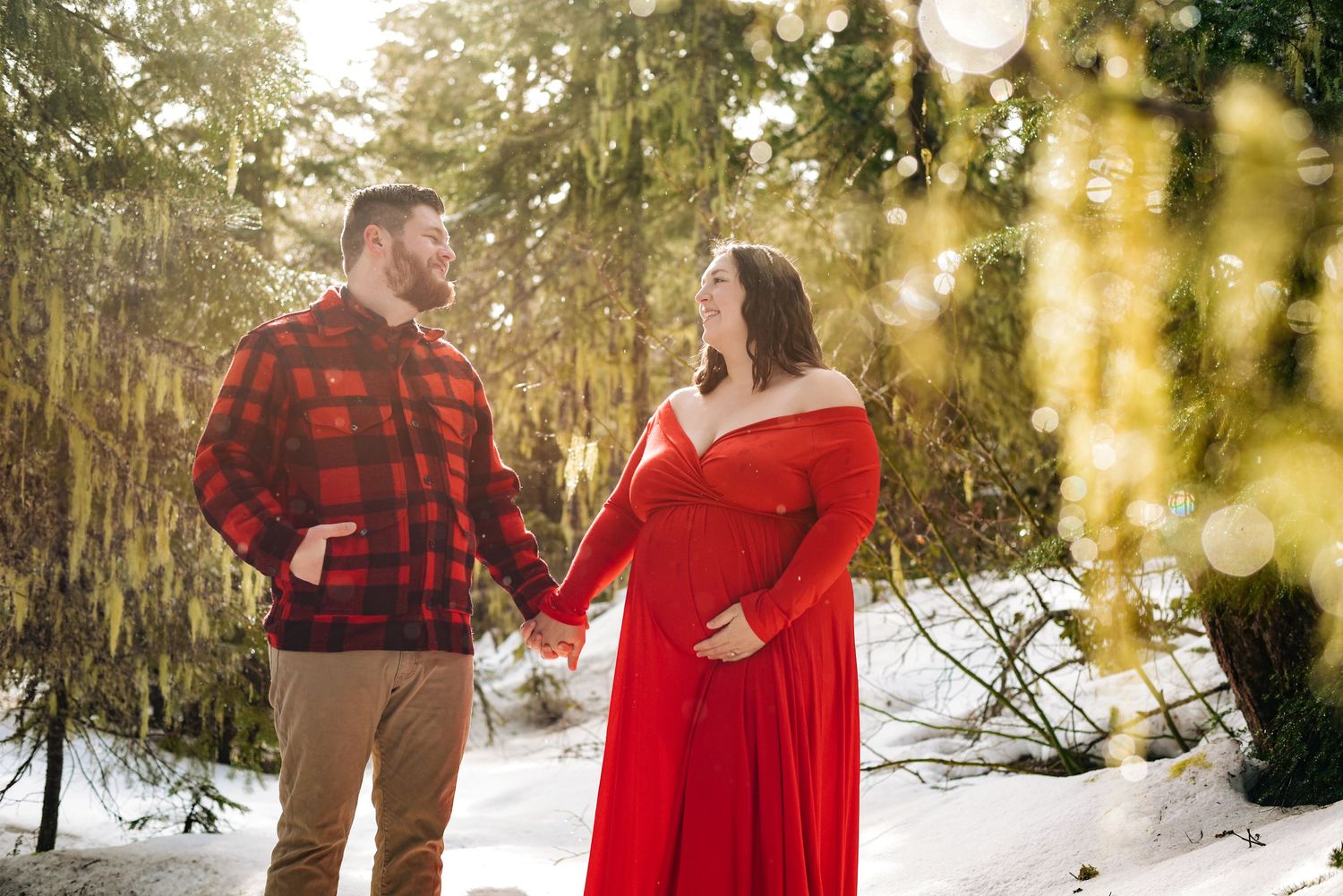 Romantic winter photoshoot with red dress and plaid shirt among snow-covered evergreens.