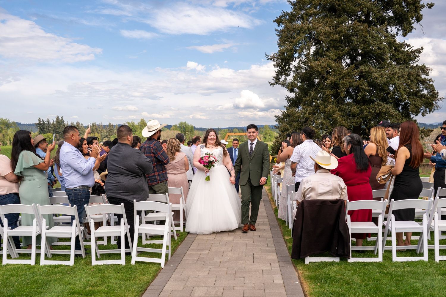Outdoor wedding ceremony with bride and groom walking down the aisle surrounded by guests on white chairs under a blue sky.