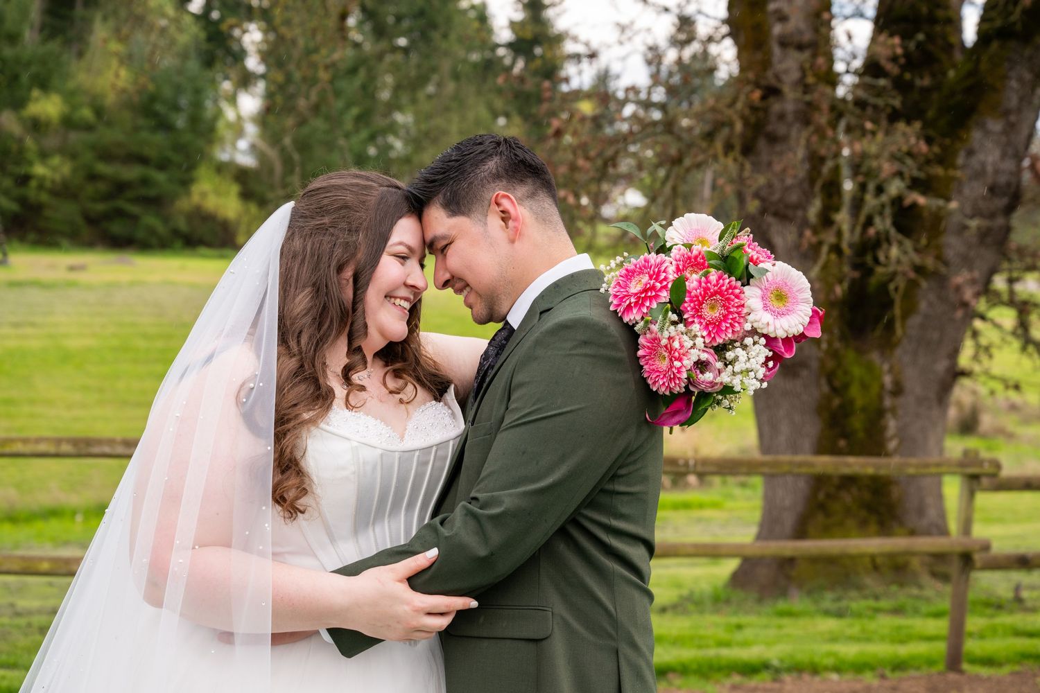 Bride and groom embracing outdoors, she holds pink gerbera bouquet, wearing white gown with veil, he in olive suit.