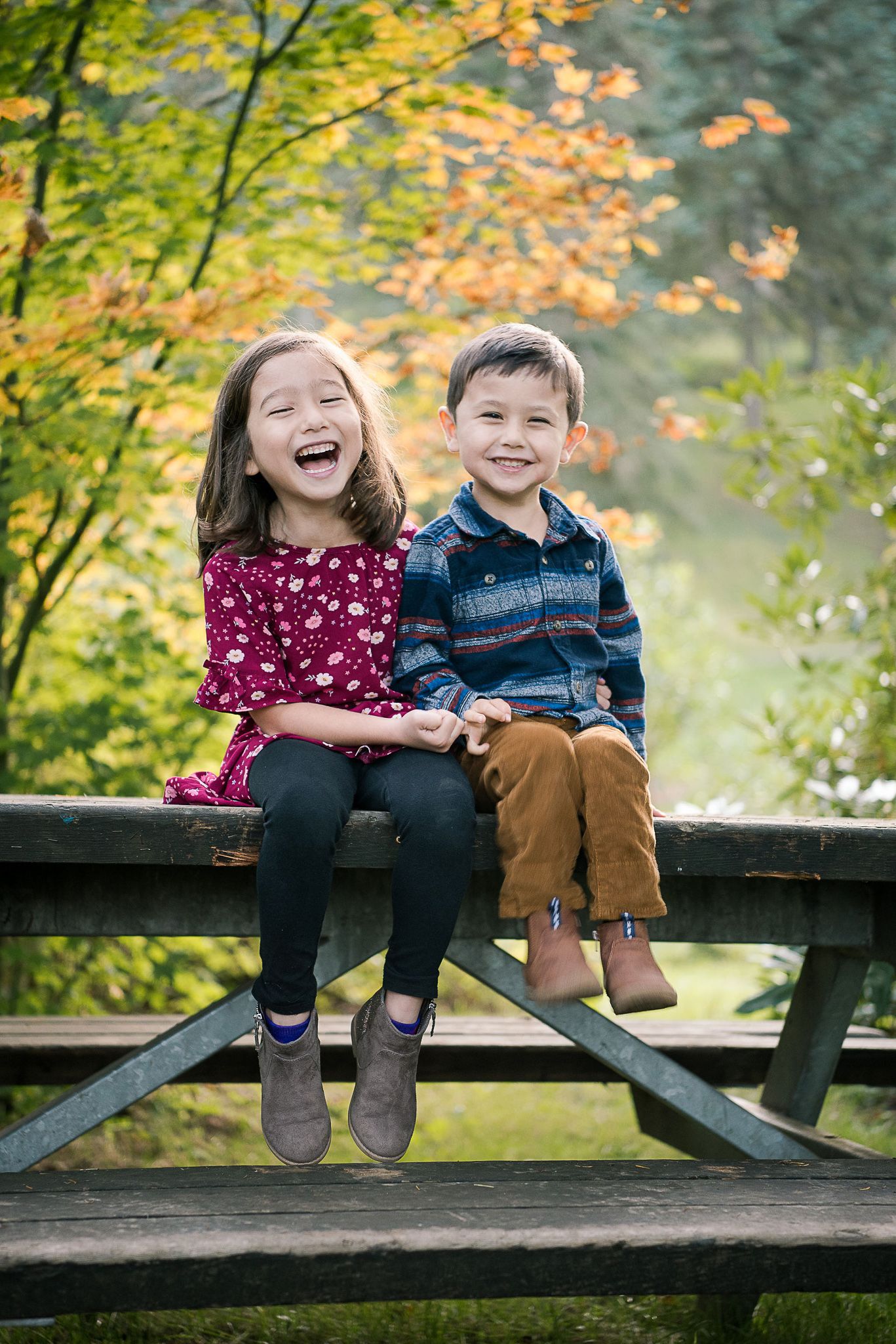 Two children sit and laugh together on a wooden fence during autumn.