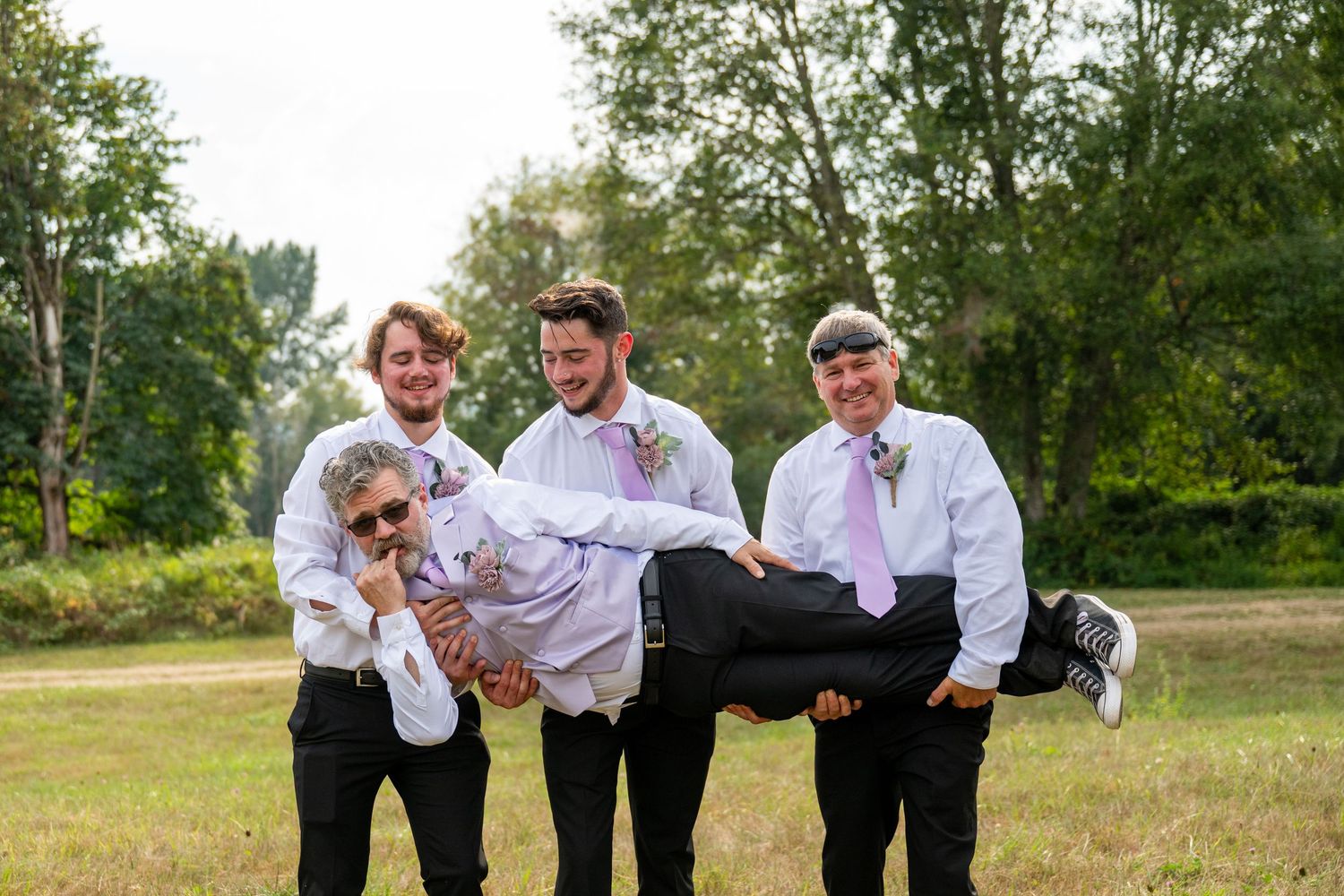 Groomsmen playfully carry groom in outdoor field during fun wedding party photo session.