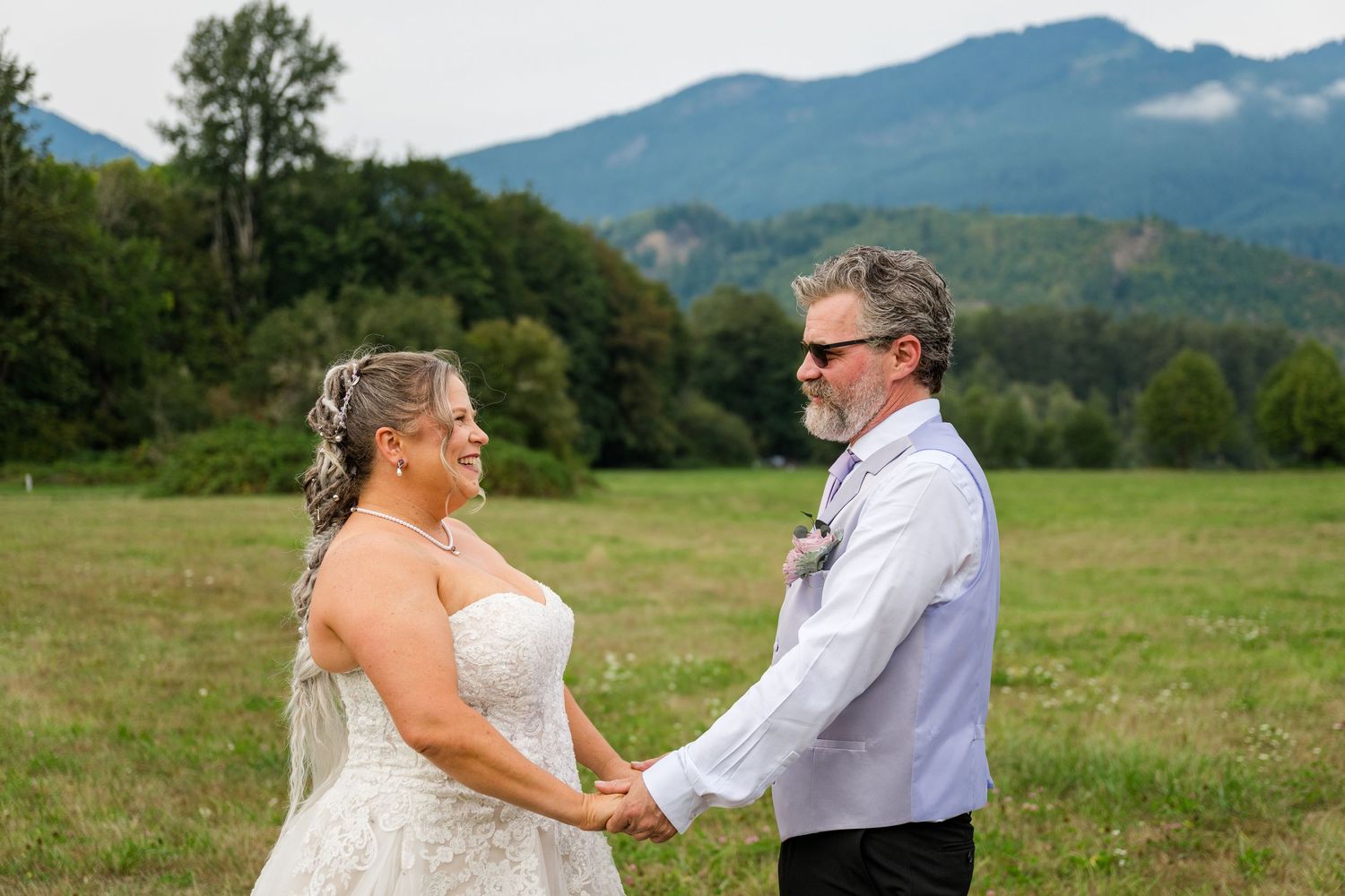 Bride and groom hold hands smiling in a green meadow with mountain backdrop during wedding ceremony.