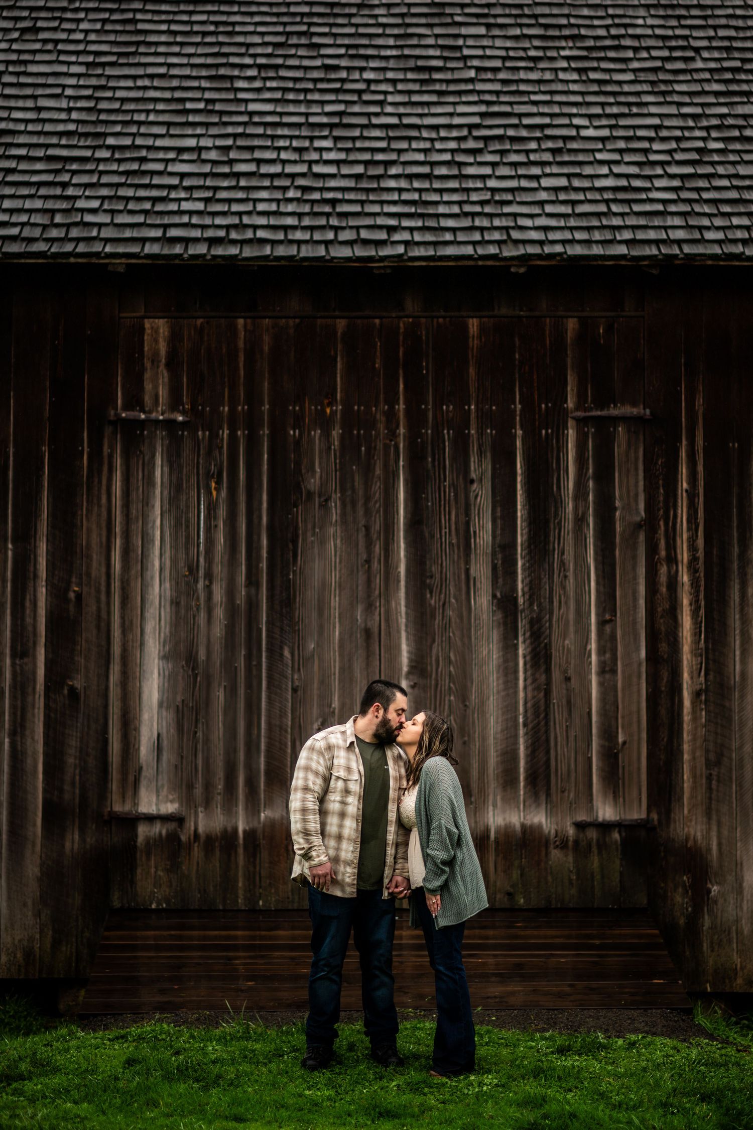 Couple sharing a tender kiss in front of a rustic wooden barn during an outdoor engagement photo session.