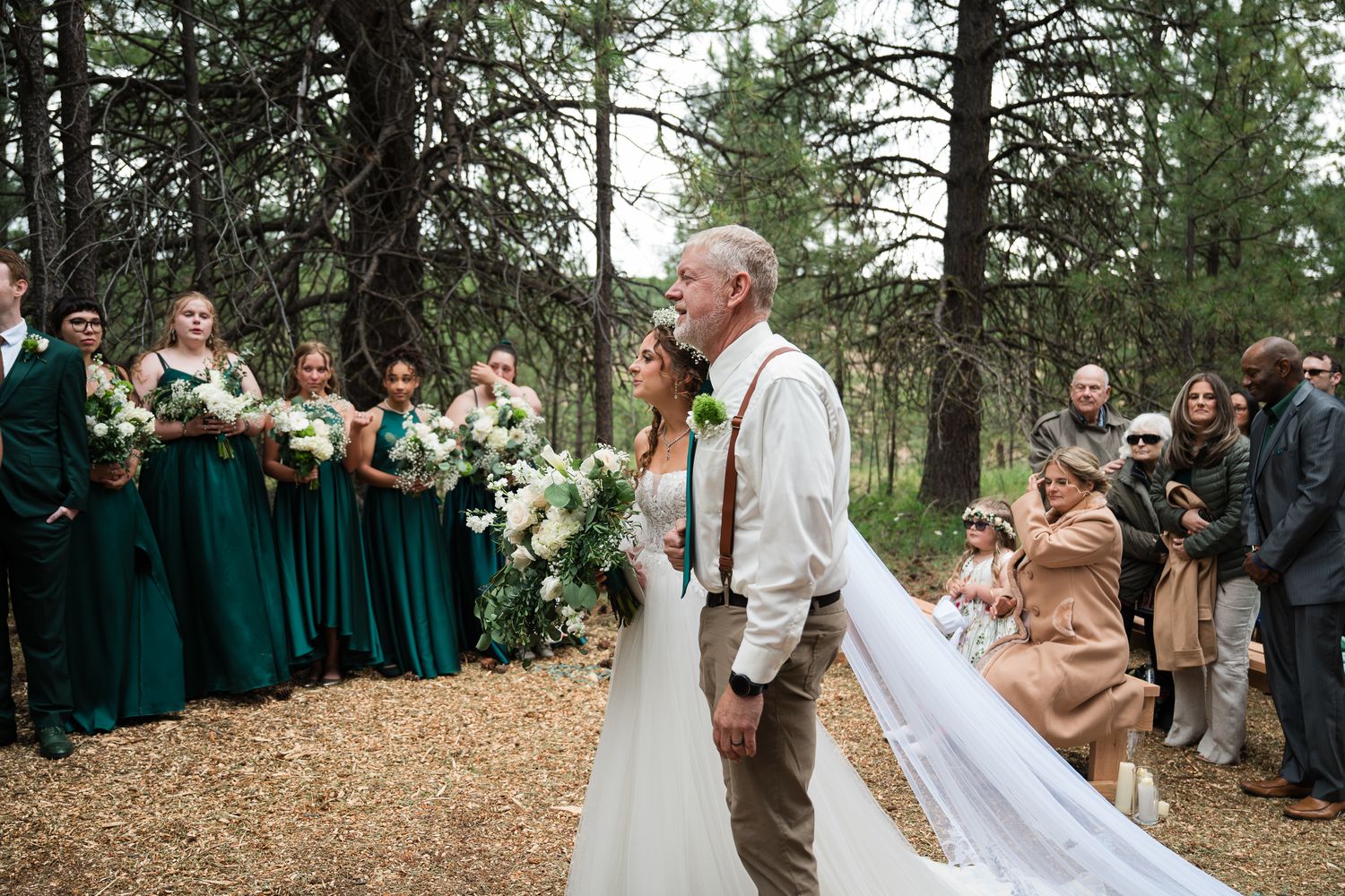 Father walks bride down forest aisle in white gown with long veil past green-dressed bridesmaids.