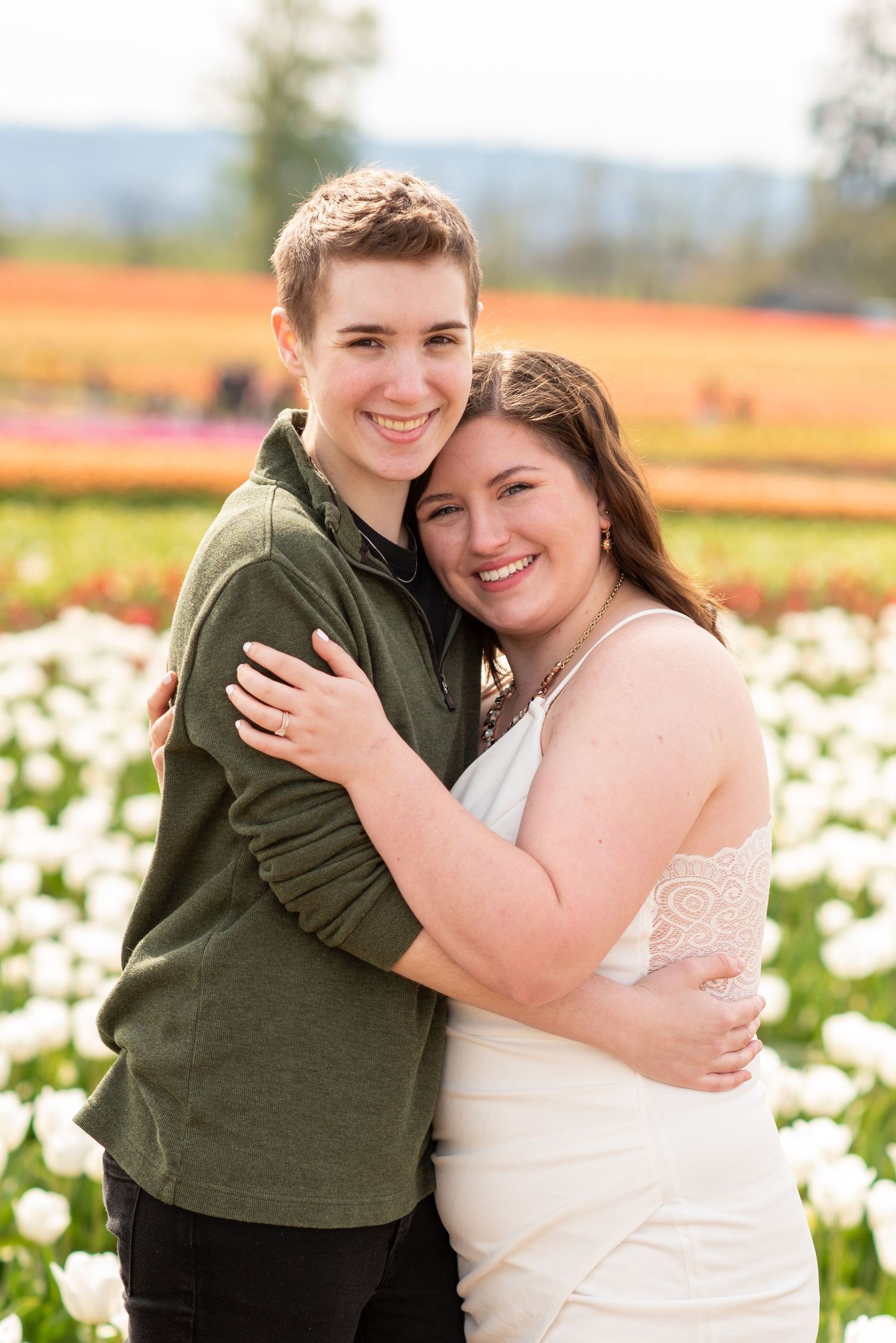 Couple embracing in field of white flowers with mountains in background.