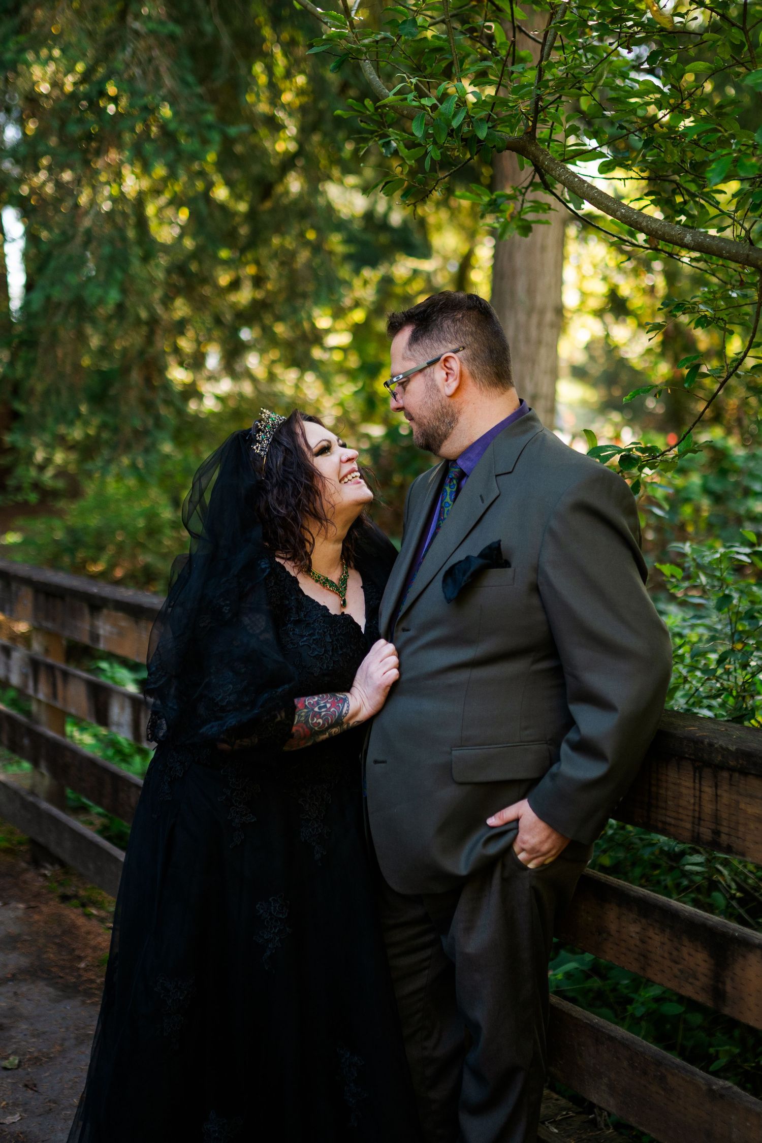 A romantic couple in dark attire share an intimate moment on a wooden bridge surrounded by lush greenery.