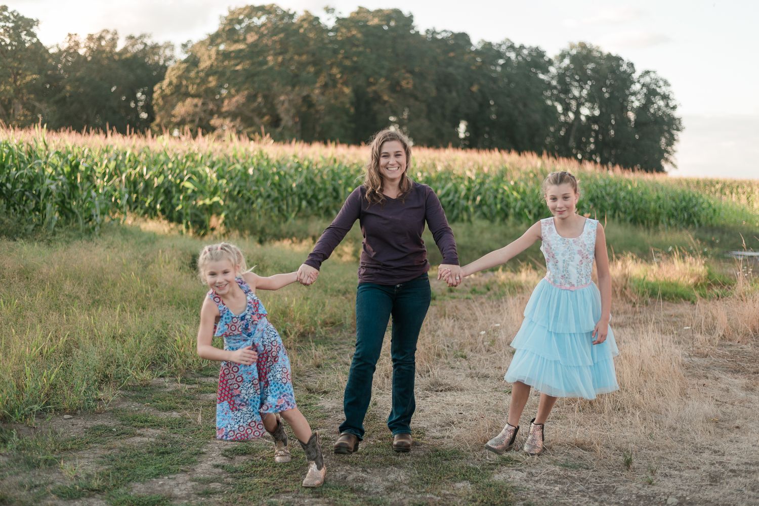 Mother holding hands with two young girls in colorful dresses near a cornfield during outdoor family portrait.