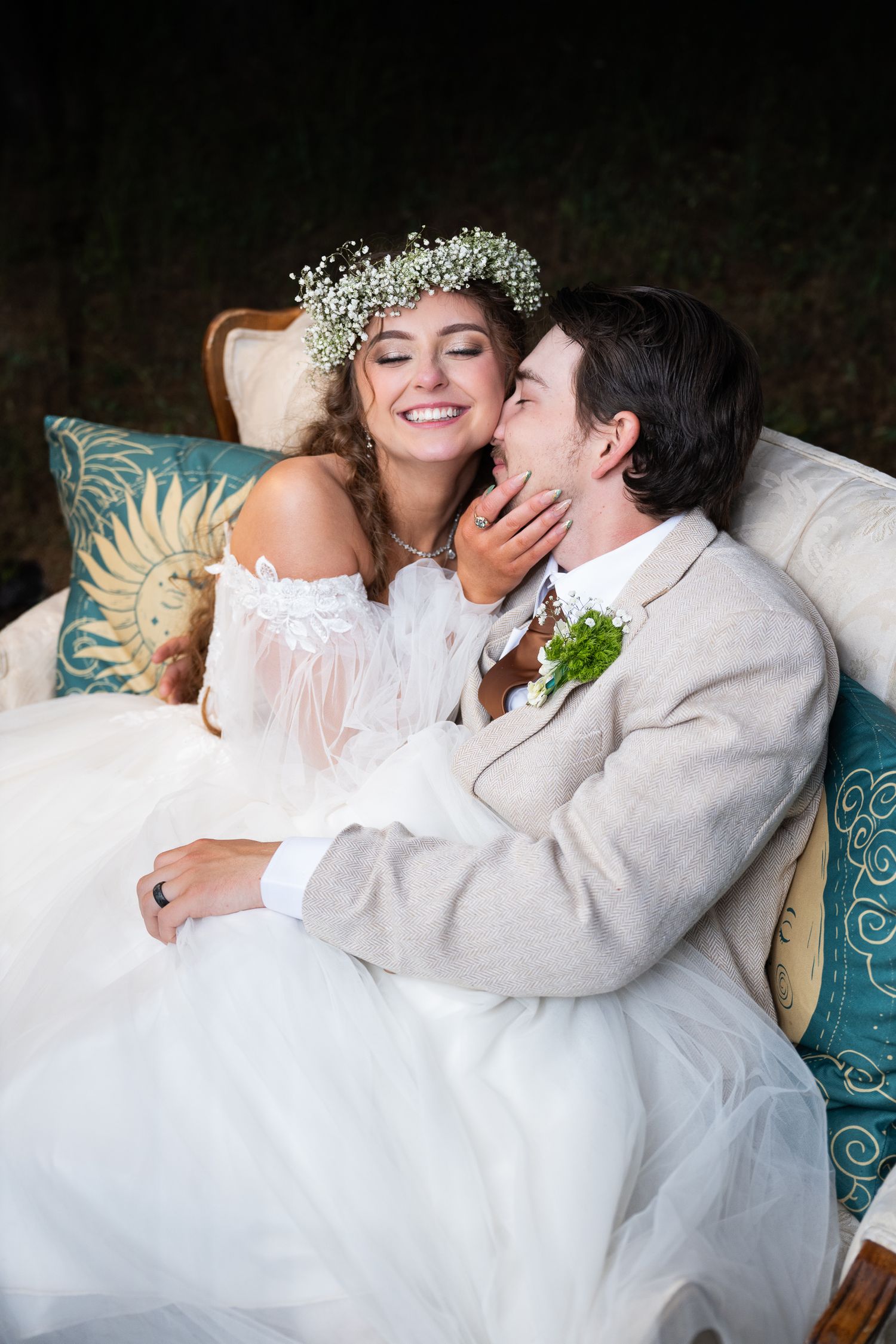 Smiling bride in floral crown and white gown sits with groom on a vintage sofa as he kisses her cheek.