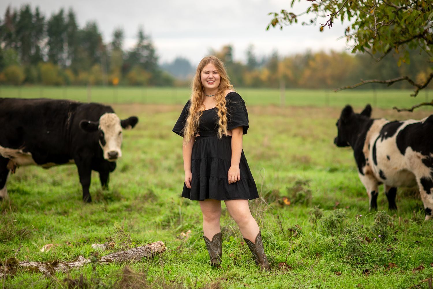 Smiling young woman in black dress poses confidently among black and white cows in a green pasture.