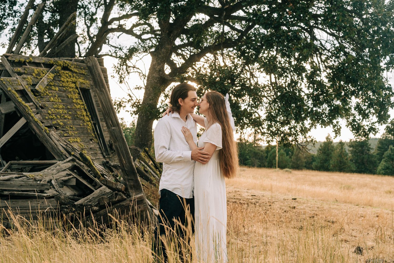A couple in white clothing posing romantically near an old wooden structure in a golden field.