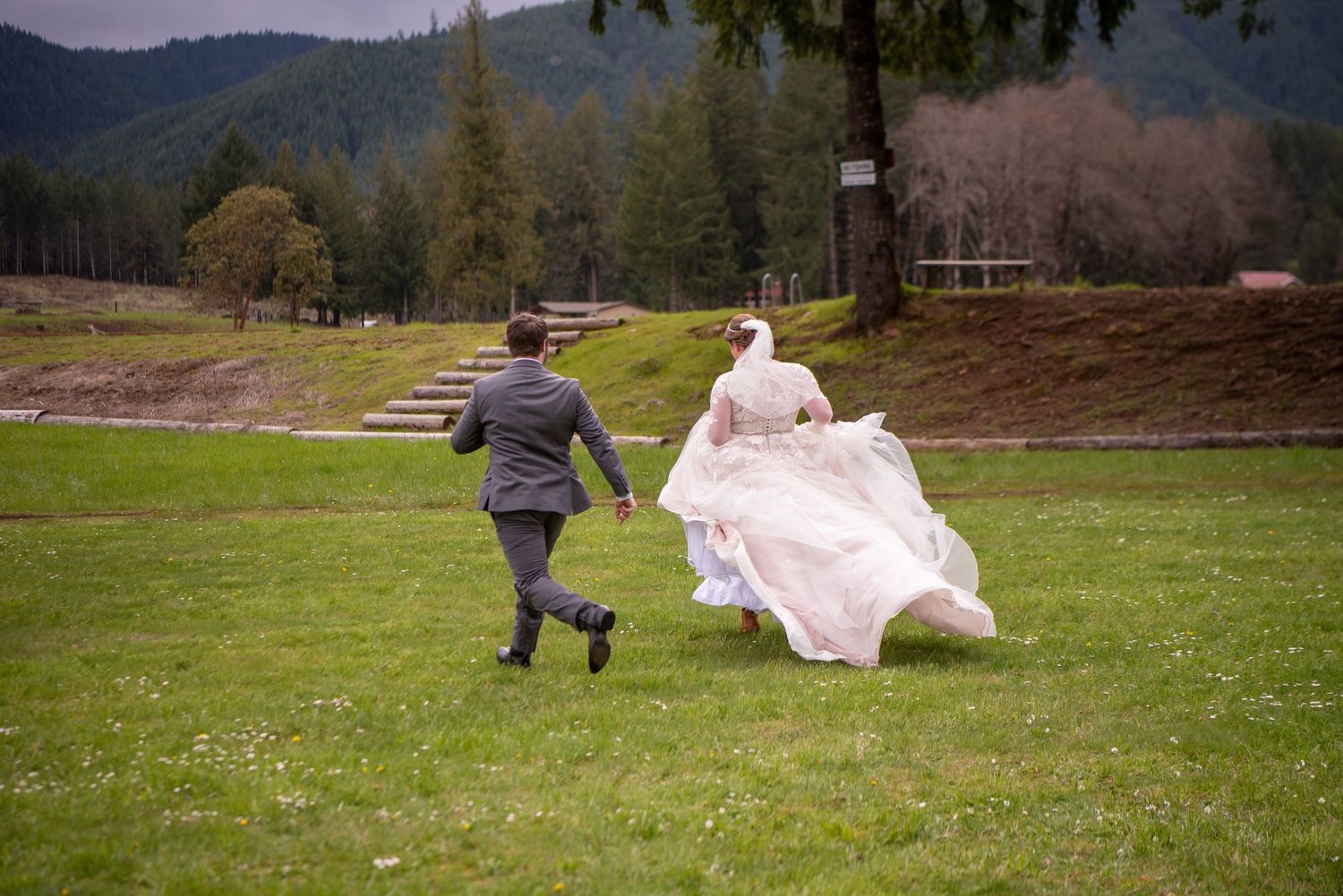 Couple runs playfully through a green field with mountains in the background.