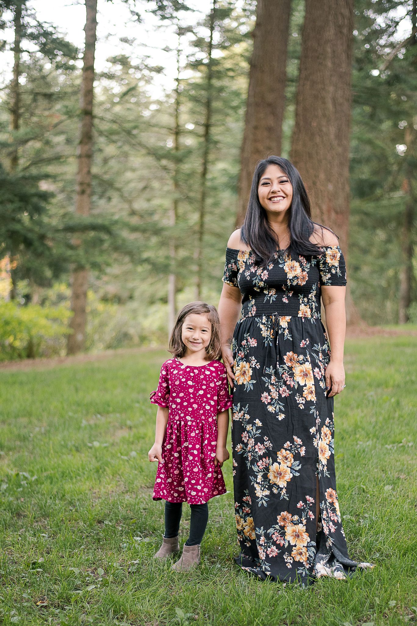 Mother and daughter in floral and polka dot dresses pose together in a sunlit forest setting.