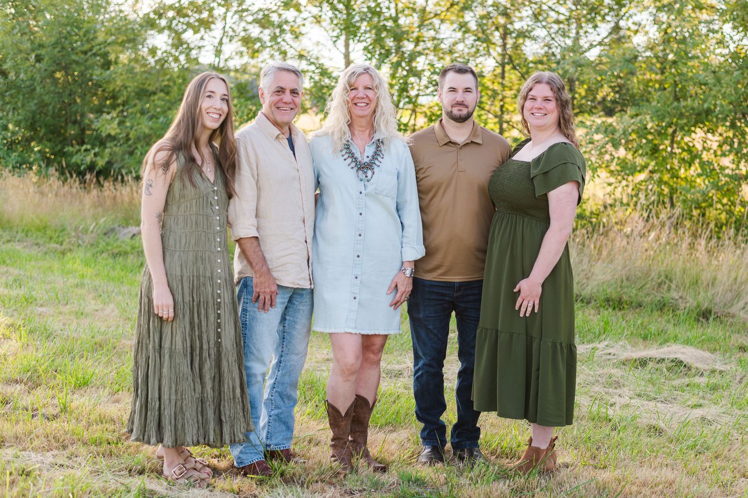 Family of five smiling together in a golden field surrounded by green trees during an outdoor portrait session.
