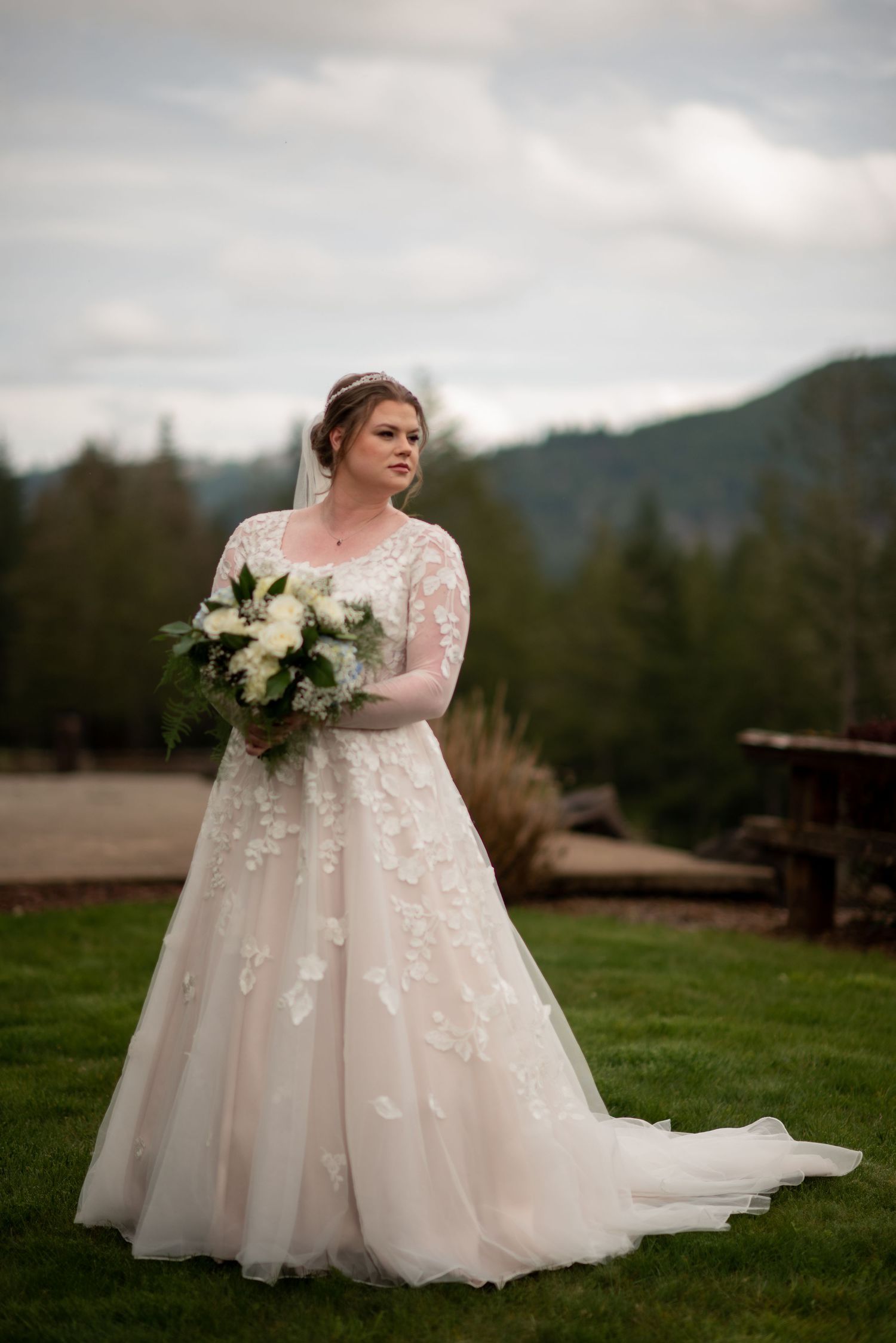 Elegant bride in a lace ball gown holding a white bouquet with mountain and forest backdrop on wedding day.