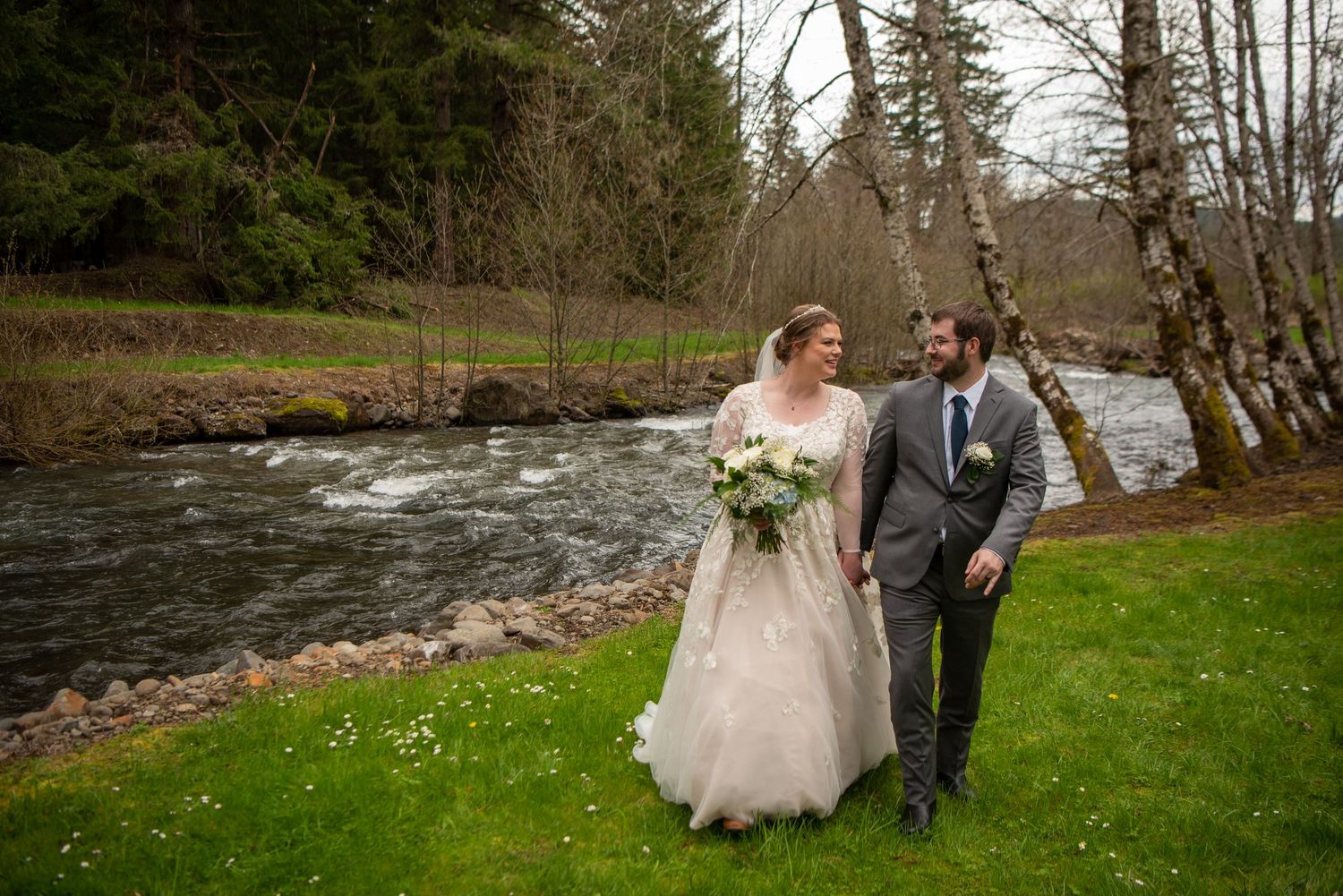 Wedding couple poses by a flowing river surrounded by trees and natural scenery.