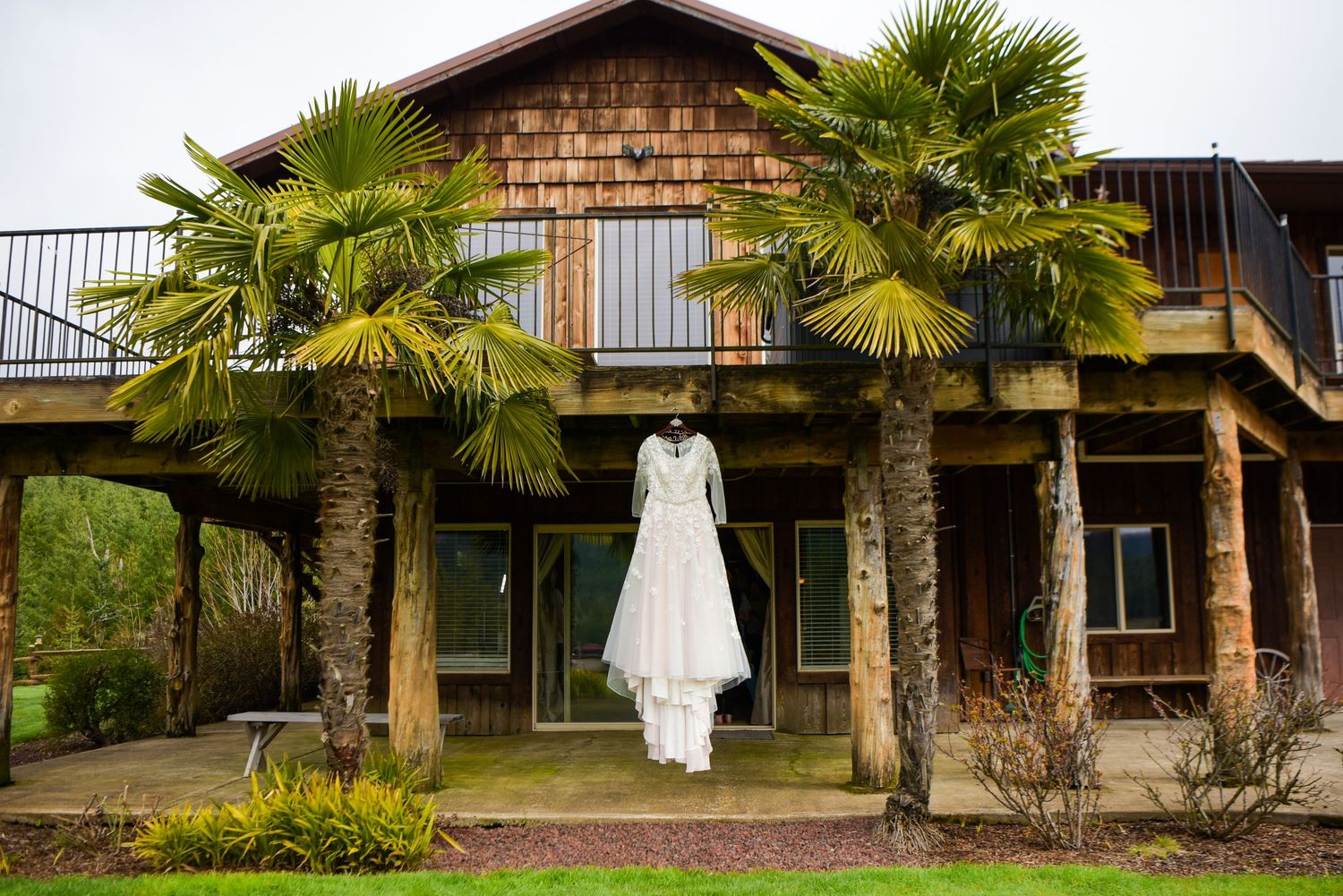 White wedding dress hanging from palm trees outside a rustic wooden venue.