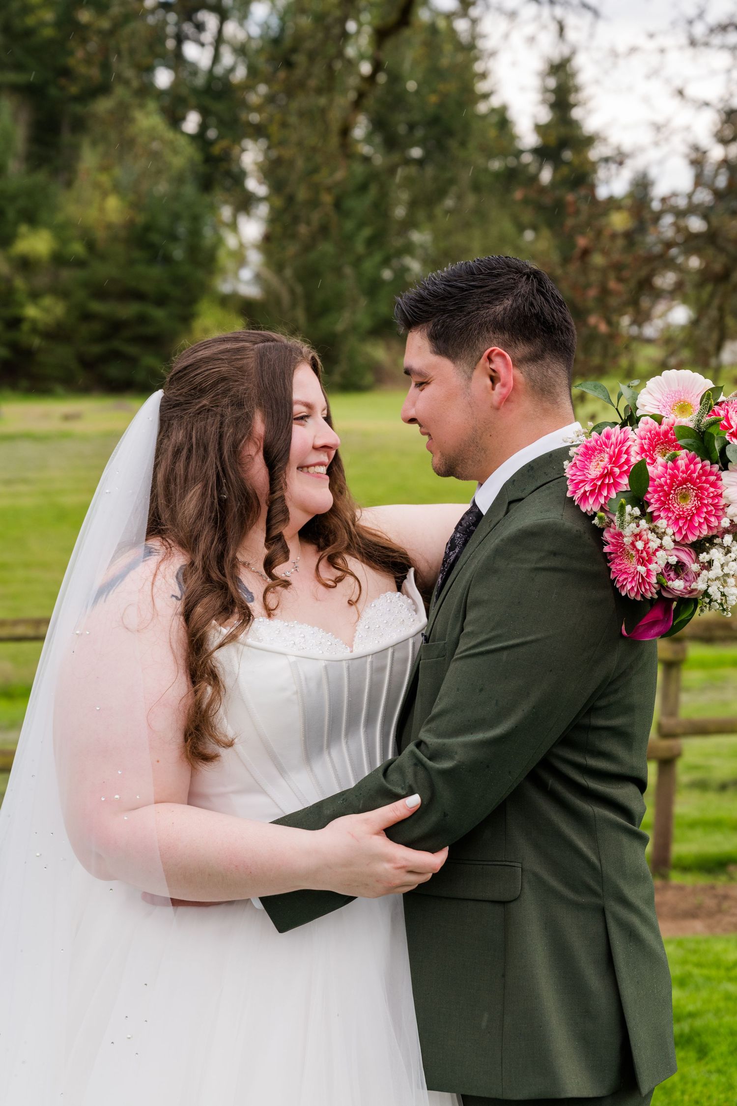 Bride in white gown and groom in green suit embracing while holding pink flowers during outdoor wedding portrait.