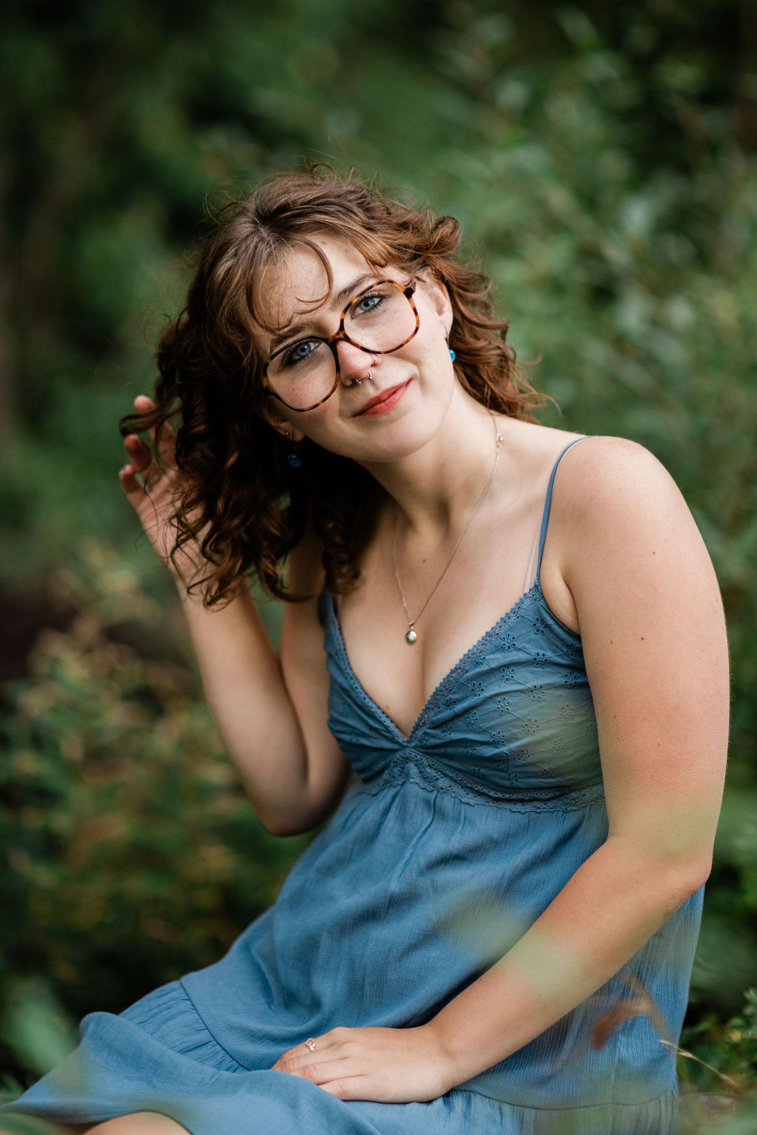 Curly-haired woman in blue dress and glasses posing outdoors among green foliage.