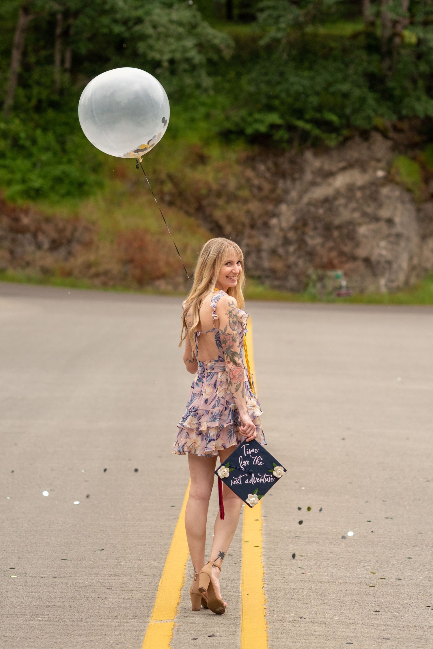Blonde graduate in floral dress holding a silver balloon and graduation cap on a scenic road.