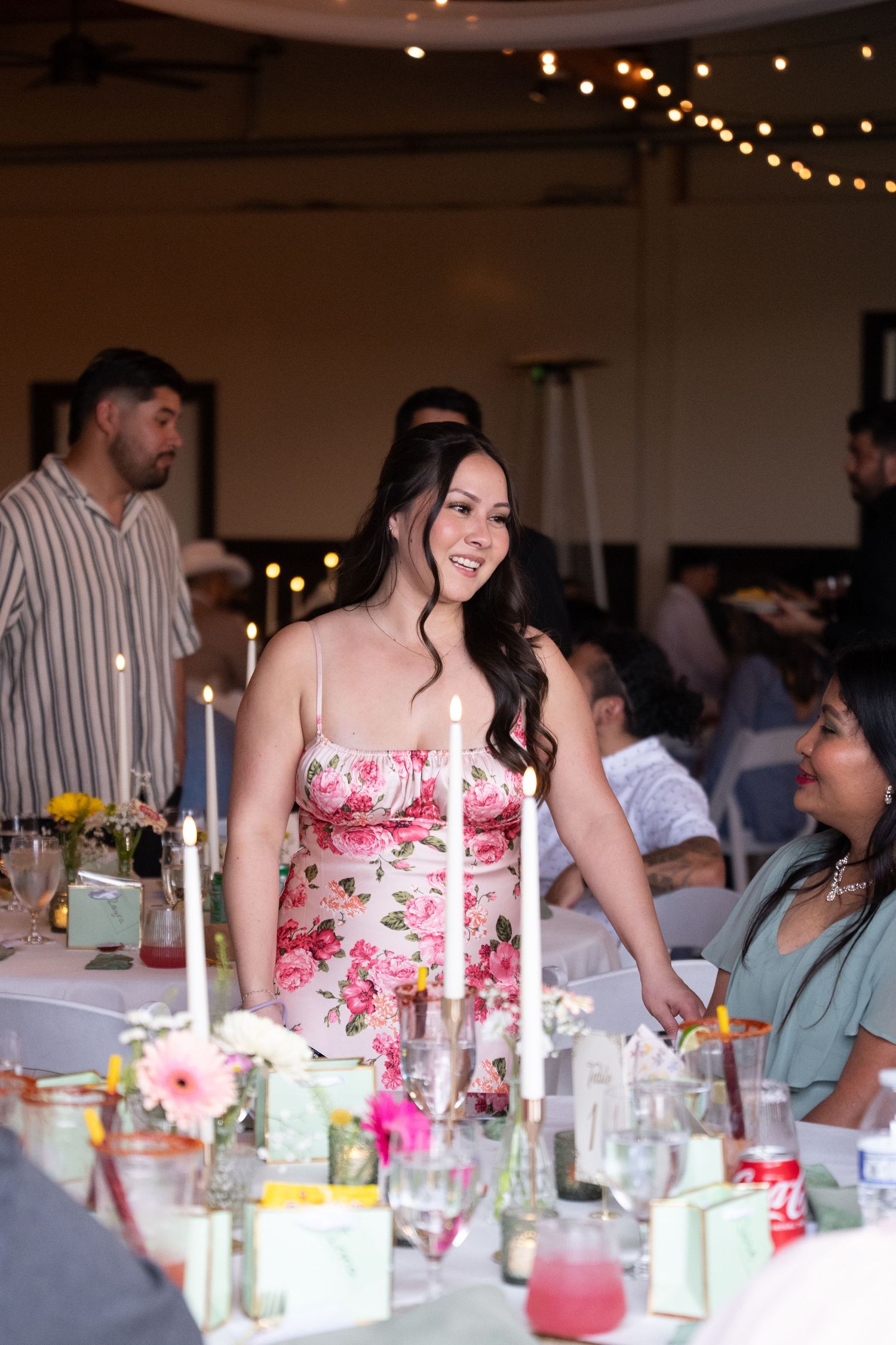 Woman in floral dress smiling at elegant wedding reception with candles and string lights.
