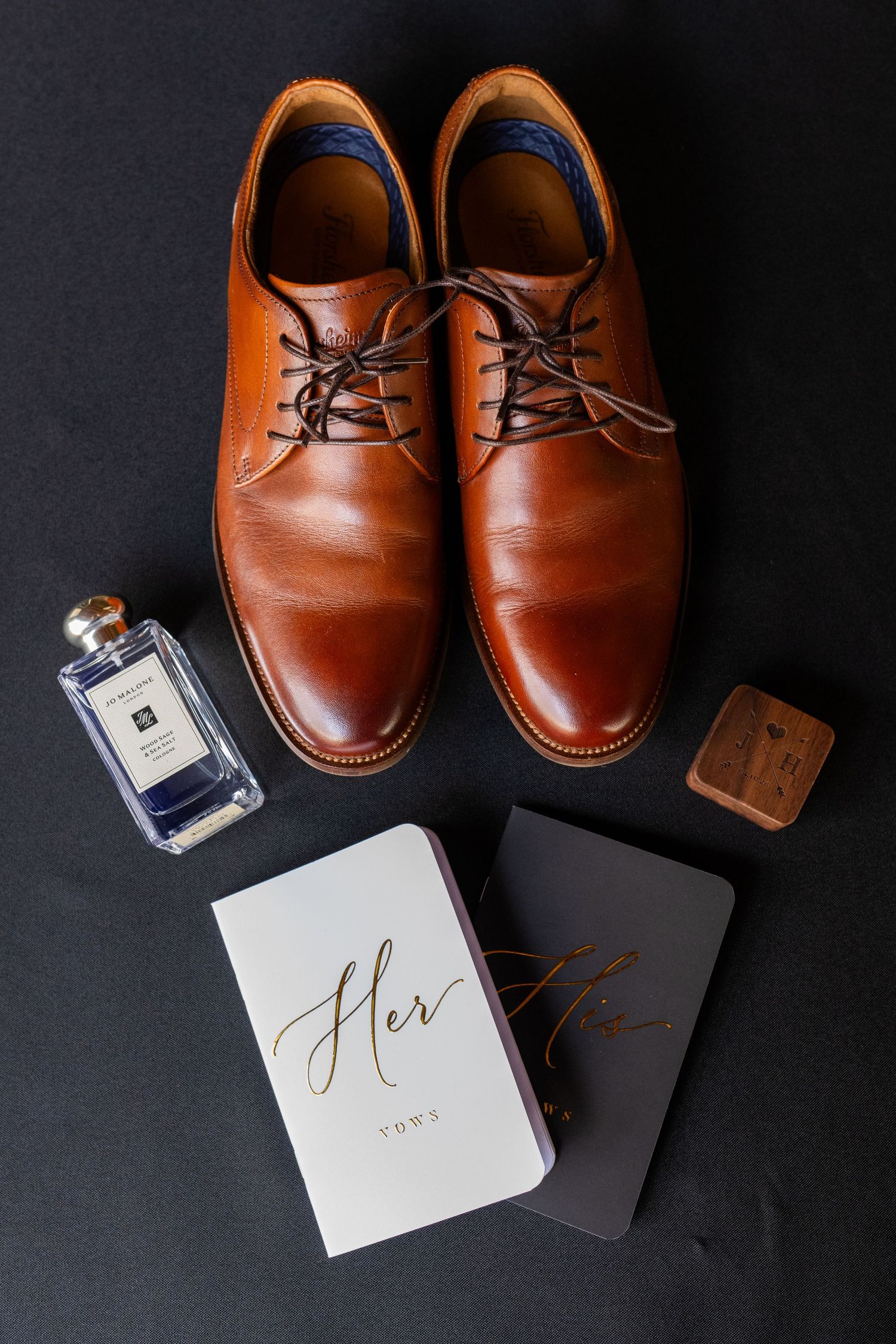 Brown leather dress shoes with cologne, vow books, and shoe polish arranged on dark background for wedding day groom details.