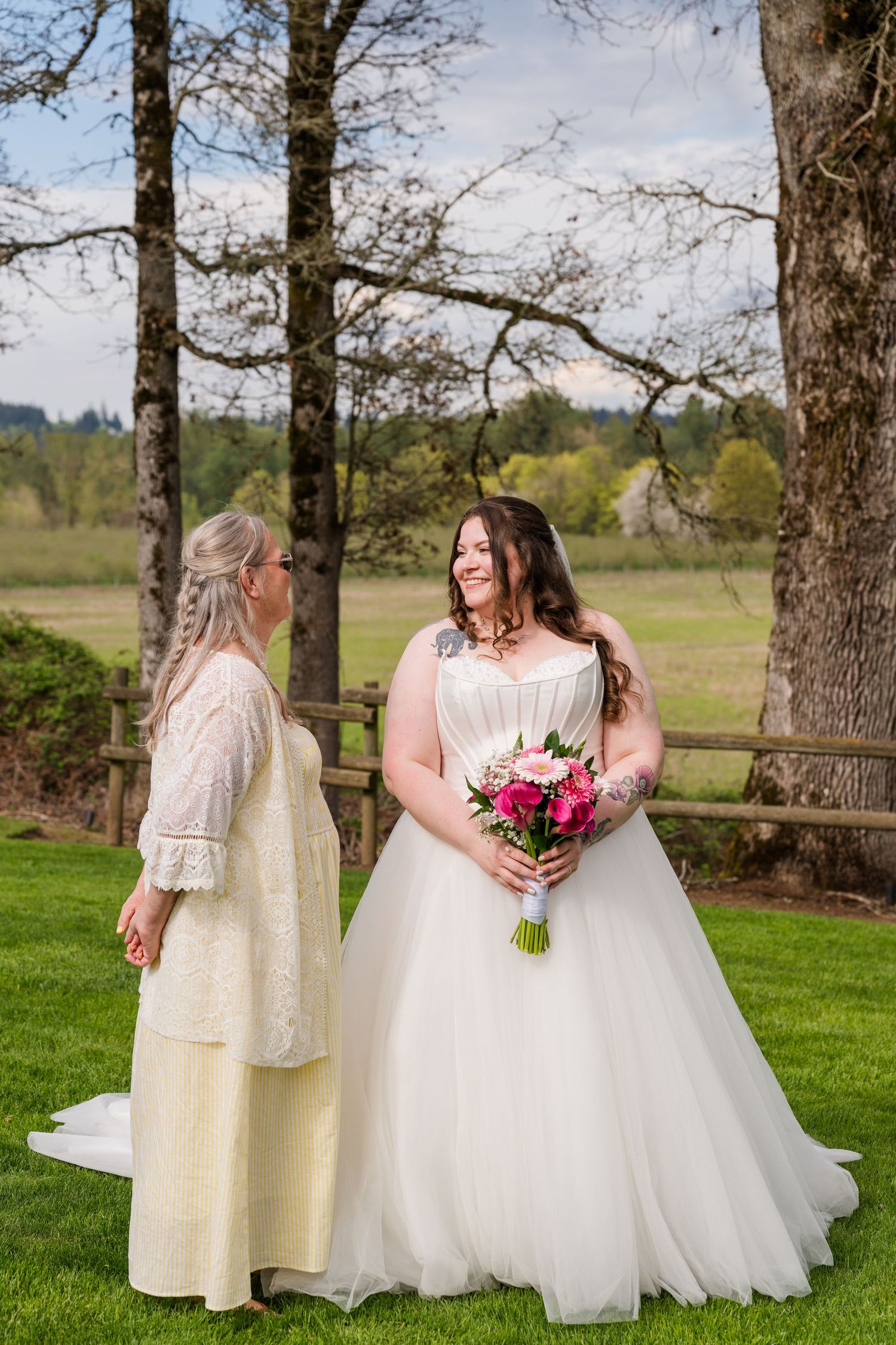Bride in white ball gown holds pink bouquet while sharing a tender moment with her mother in a lace dress outdoors.