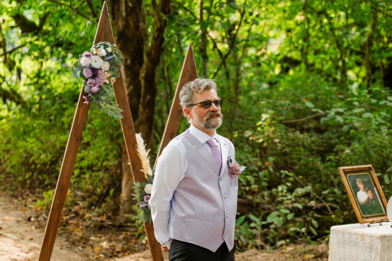 Groom in lavender vest and sunglasses stands beside wooden triangular arch decorated with flowers in forest.