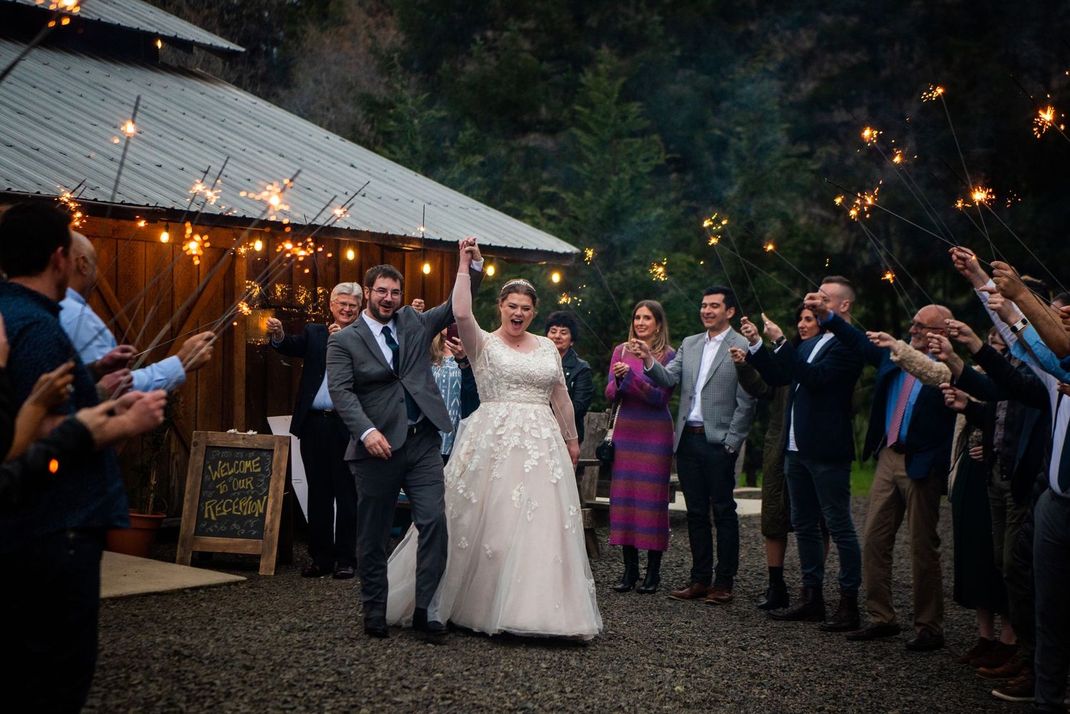 Joyful bride and groom exit their rustic barn wedding reception through a glowing sparkler send-off with guests.