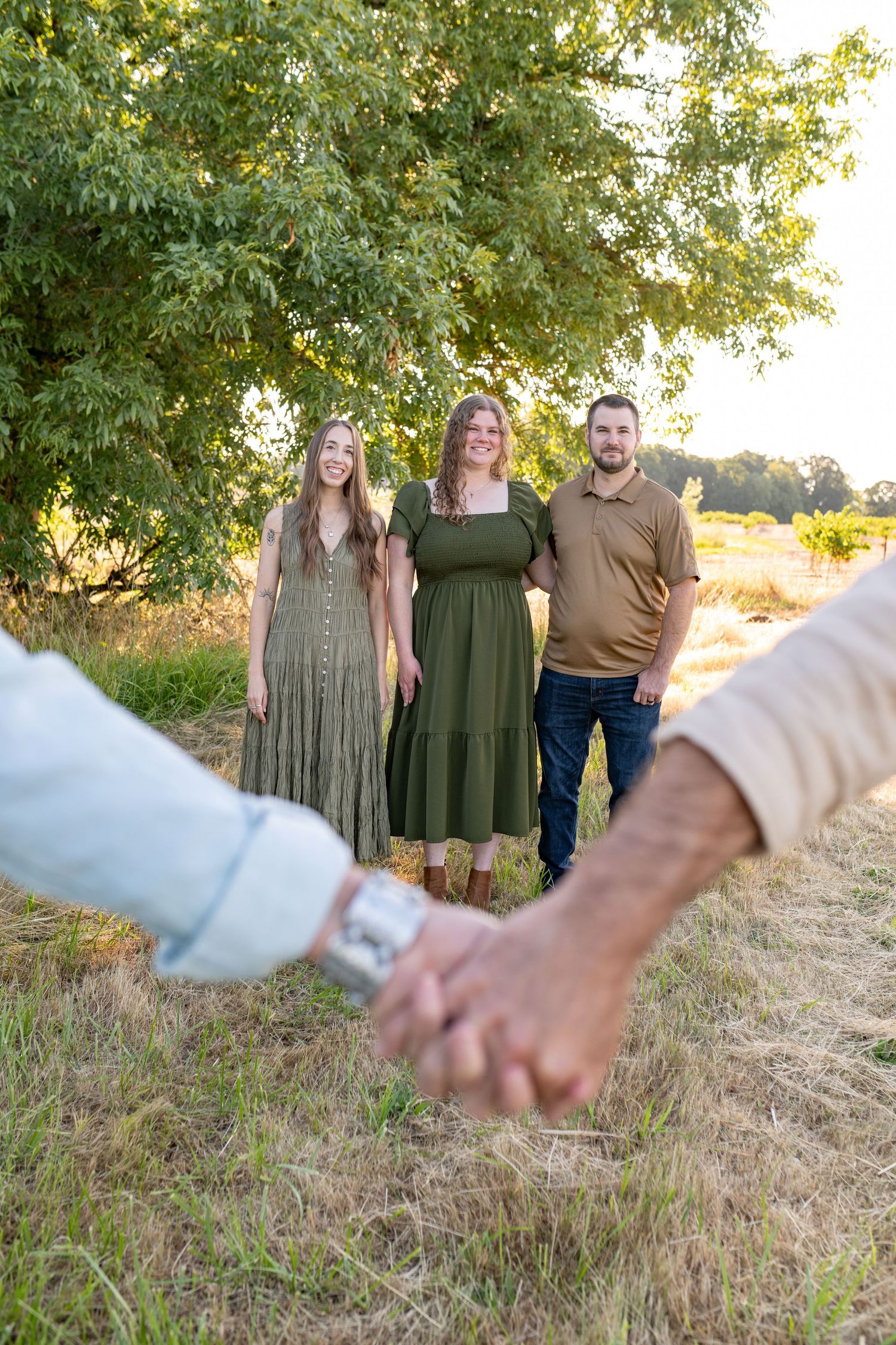 Three adult siblings standing under a large tree while parents hold hands in the foreground during family photos.