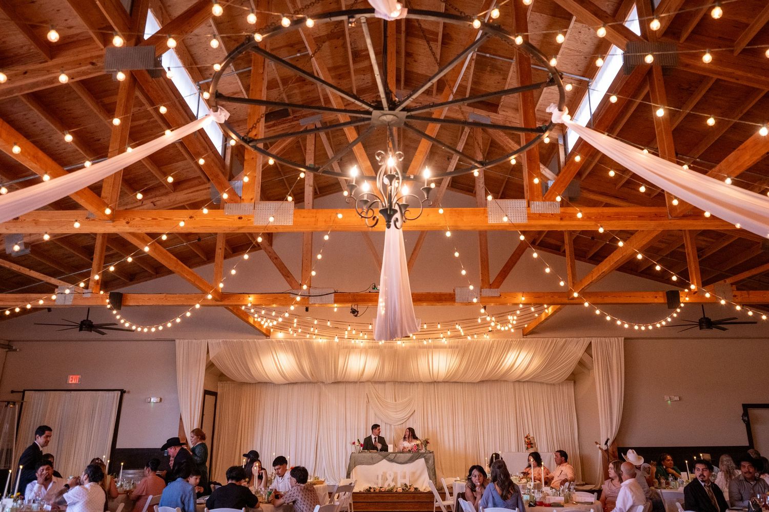 Elegant barn wedding reception with string lights, wooden ceiling, chandelier, and draped fabric decor.
