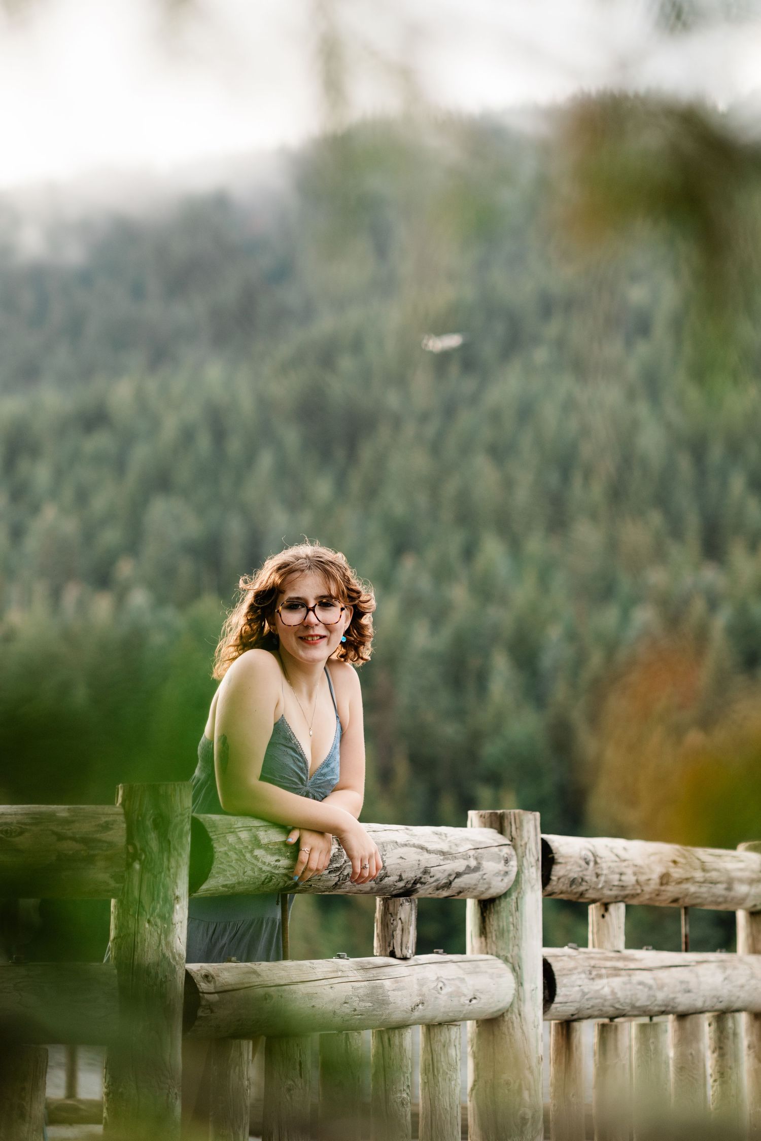 Woman in green dress leans on rustic wooden fence railing with forest mountain backdrop in soft focus.