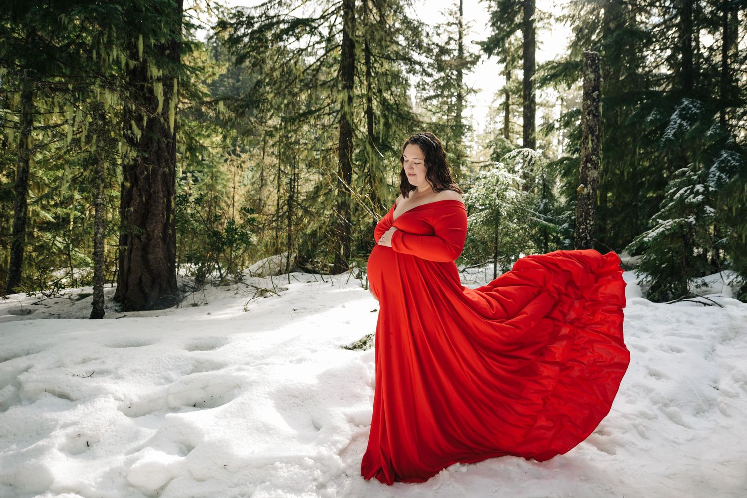 A red dress flows dramatically against white snow in an evergreen forest landscape.