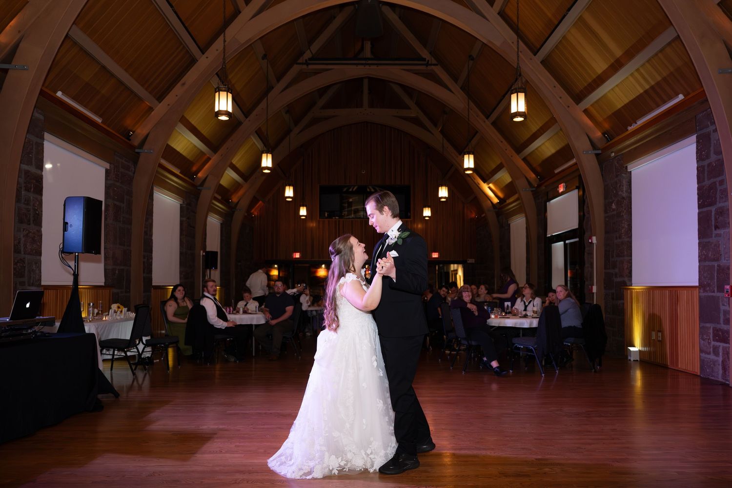 Bride and groom share first dance in elegant wood-beamed wedding reception hall with warm pendant lighting and seated guests.