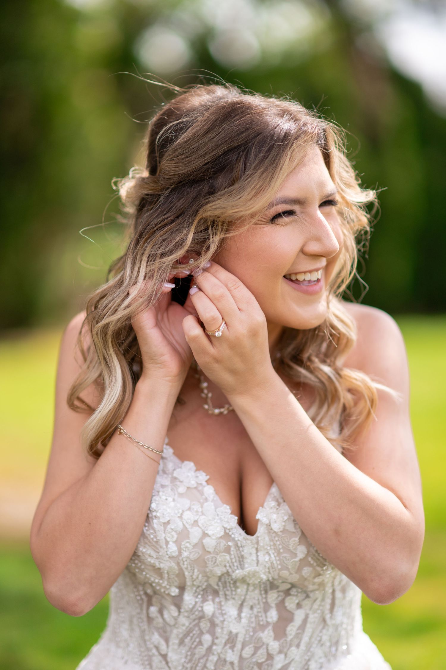 Radiant bride in lace strapless gown smiles while adjusting earring outdoors with blurred green background.