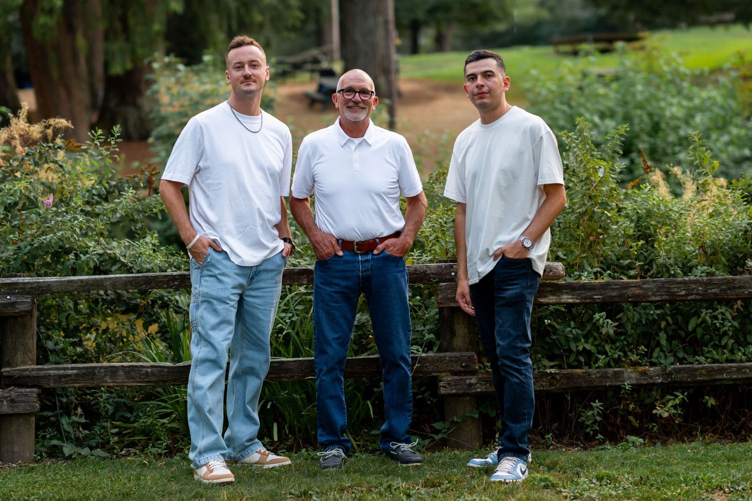 Three men in white shirts and jeans posing by a wooden fence in a garden.