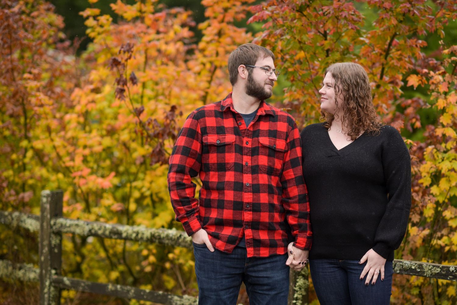 Couple posing together against vibrant autumn foliage in red plaid and black outfits.