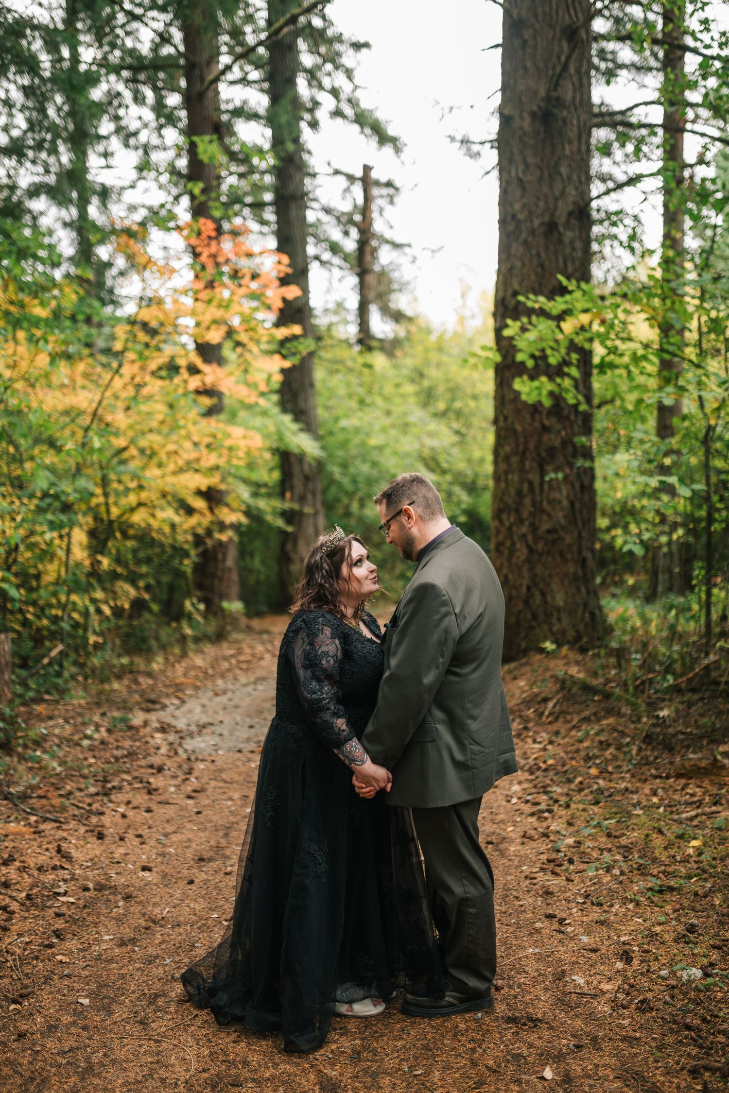 An intimate forest portrait session with a couple on a leaf-covered path surrounded by tall evergreen trees.