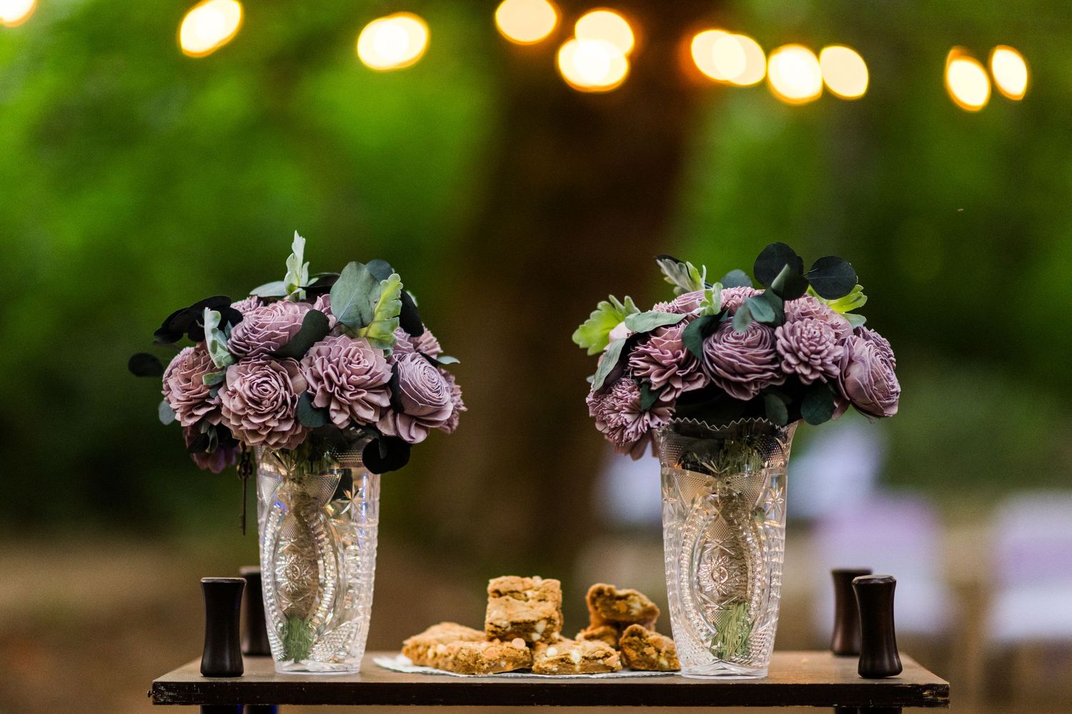 Two purple floral centerpiece arrangements with sola flowers in crystal vases displayed on wedding reception table.