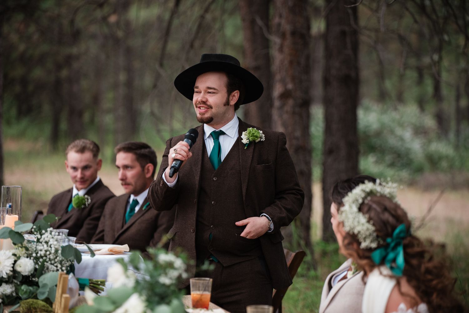 Man in brown suit and wide-brim hat gives a toast at an outdoor wedding reception in a pine forest.