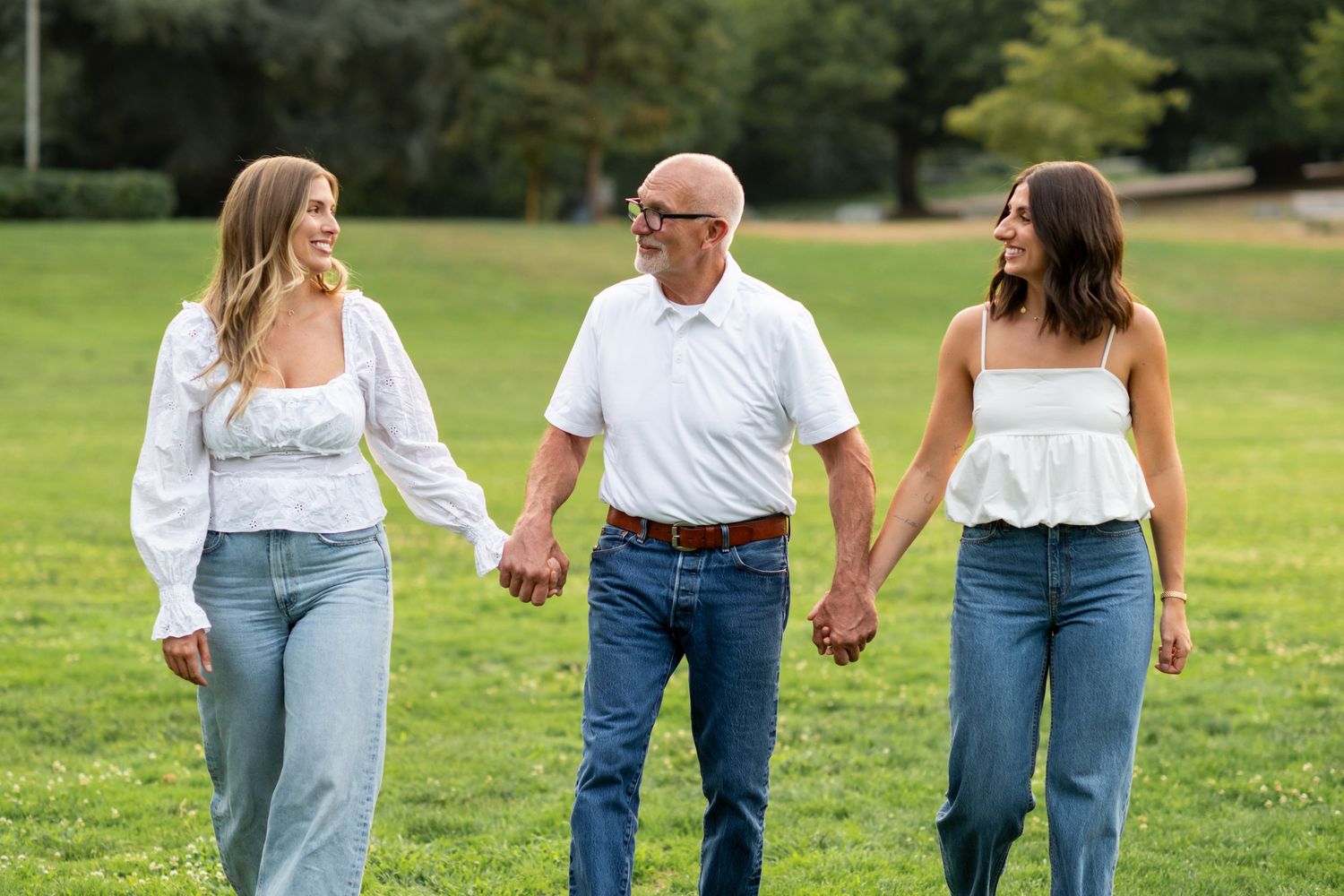Father walks hand in hand with two daughters in white tops and jeans in a park.