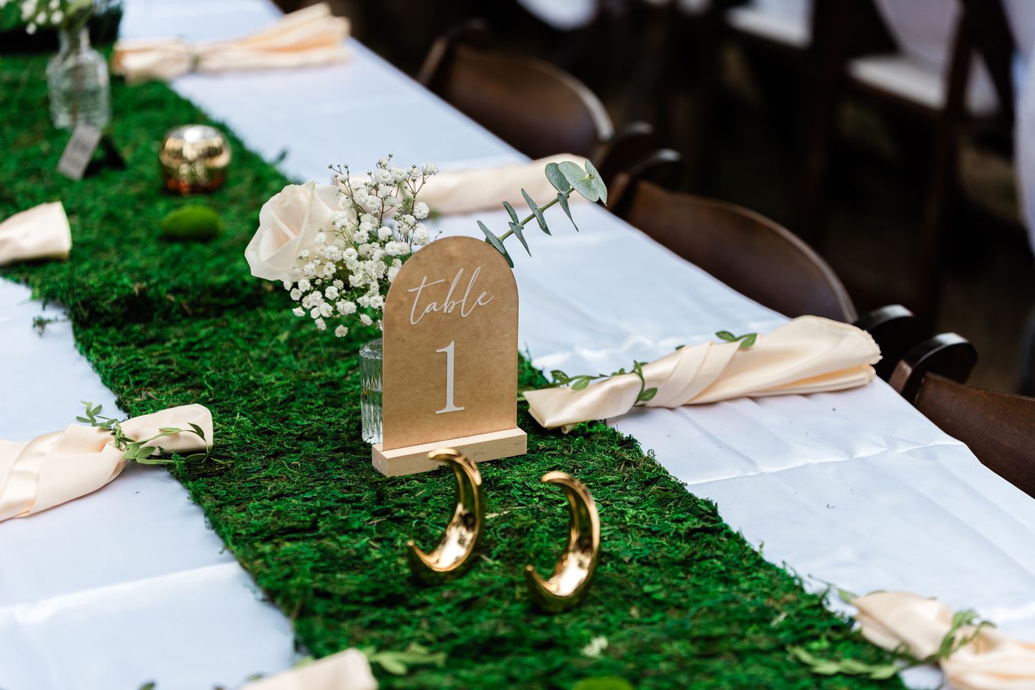 Wedding reception table with moss runner, baby's breath, blush napkins, and a gold Table 1 sign.