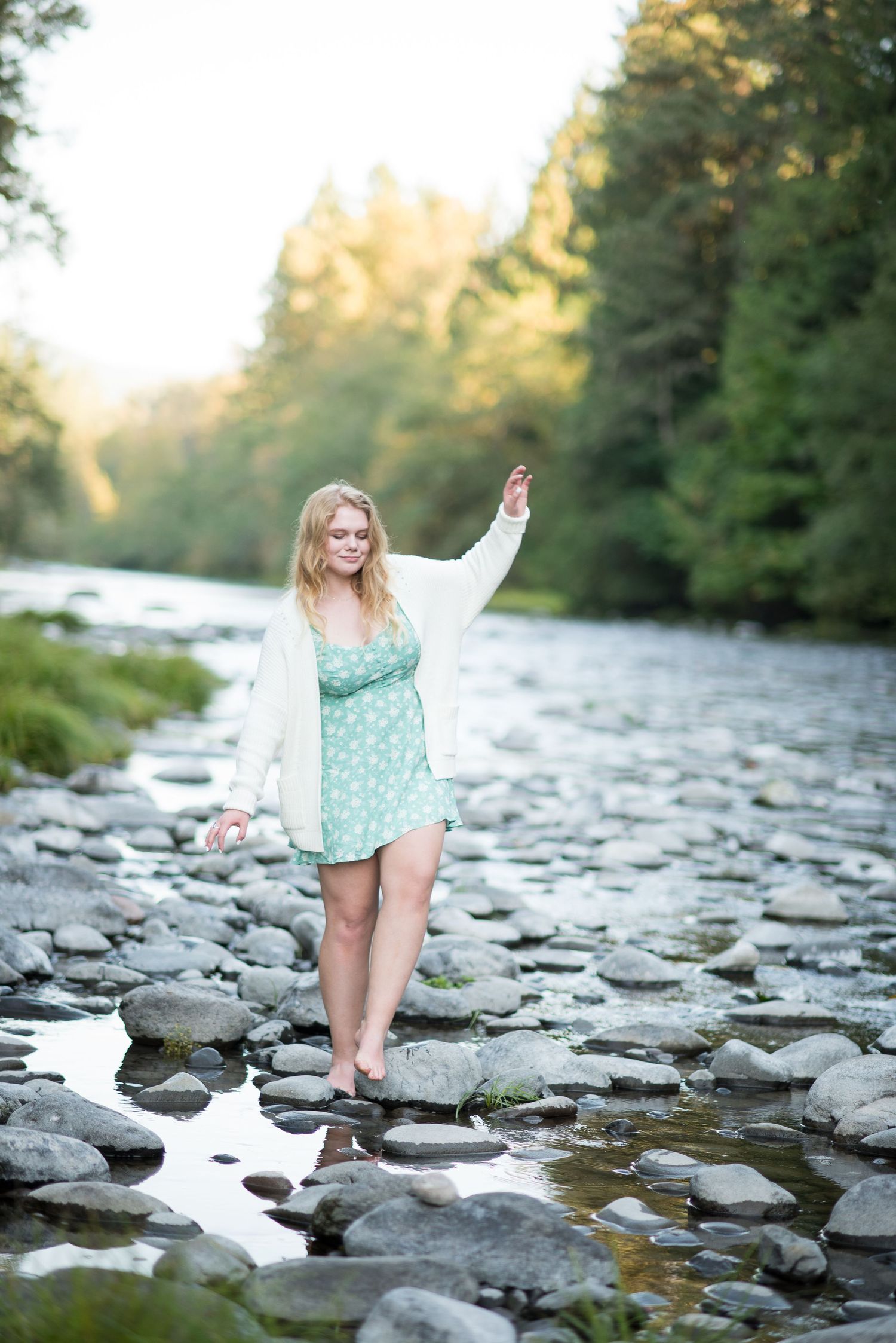 Barefoot blonde woman in green polka dot dress balancing on rocks by a peaceful river surrounded by lush trees.