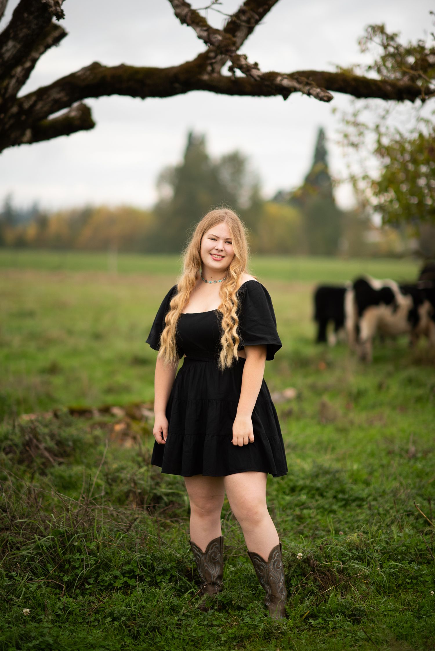 Smiling blonde teen in black dress and cowboy boots stands under bare tree branches with cattle behind.