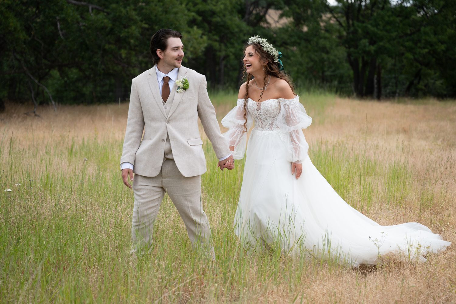 Bride and groom walk hand-in-hand through a golden meadow, she in a flowing white gown, he in a beige suit.
