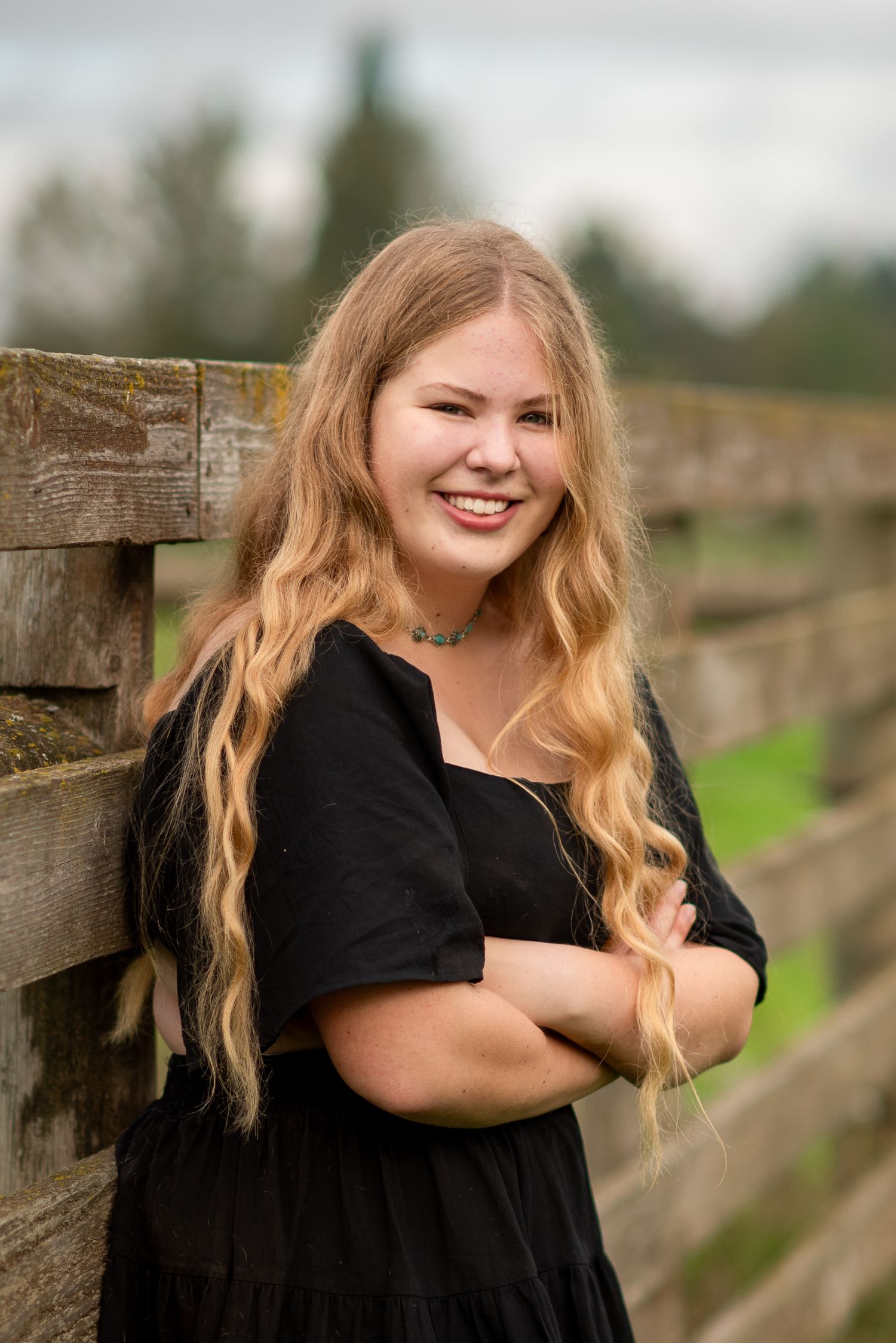 A person in black attire poses by a wooden fence in a countryside setting.