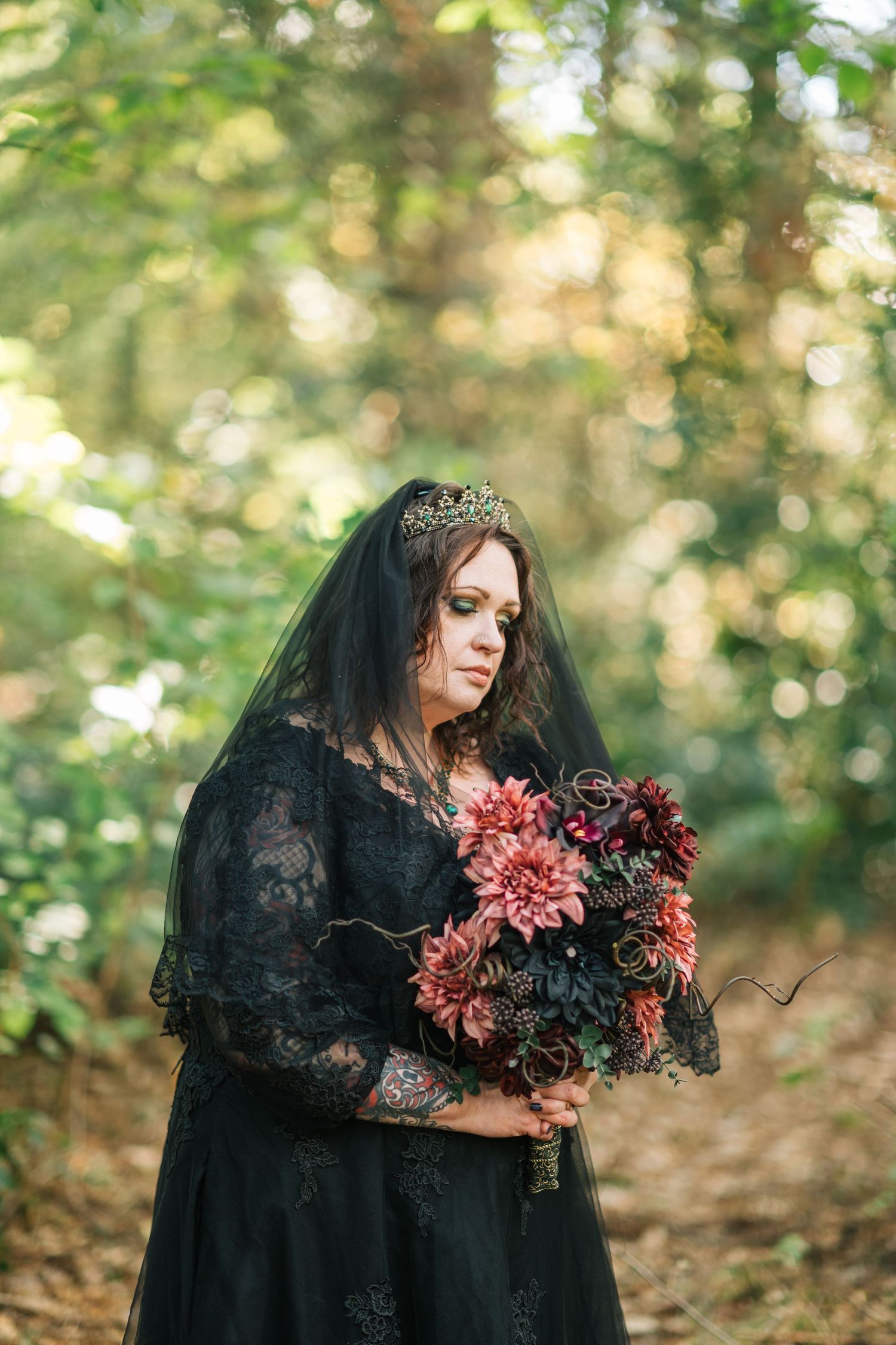 A person in a black gothic dress and veil holds dark burgundy flowers in a moody forest setting.